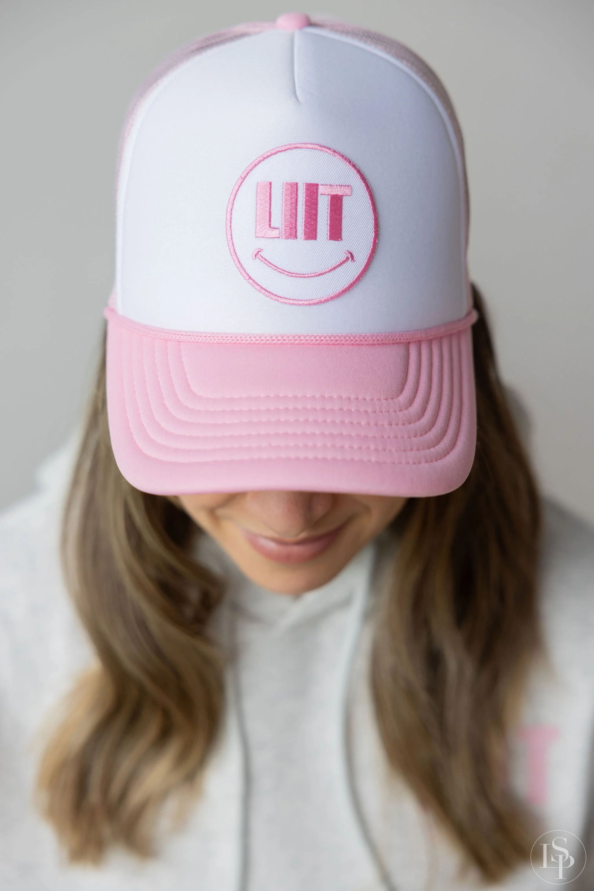 Person with long wavy hair wearing a white and pink trucker cap with a smiley face and the word 'LIT' embroidered on the front.