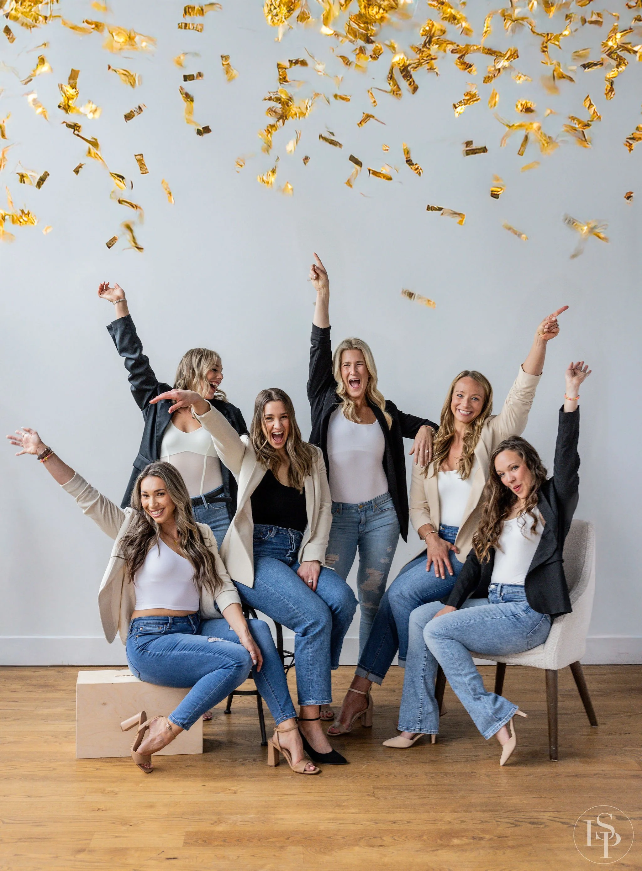Lifestyle team photographer near me in Cleveland Ohio - Group of eight women celebrating, some standing and some sitting, with confetti falling around them in a bright room with a wooden floor and white walls.