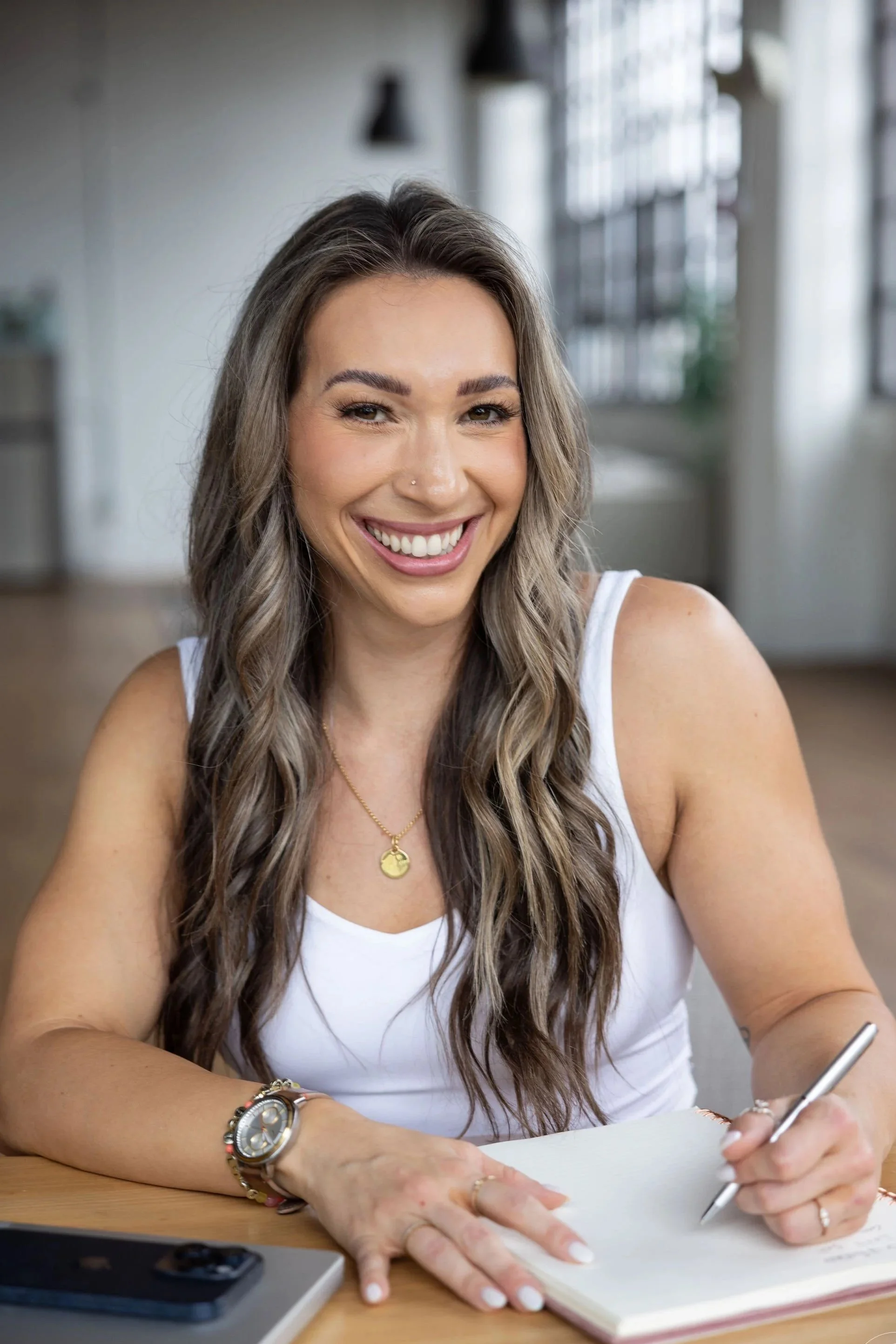 Branded photography of Smiling woman with long wavy hair, wearing a white tank top and gold necklace, writing in a notebook at a wooden table, with a smartphone and a pen nearby, in a bright room with large windows. lifestyle headshots near me -Ohio