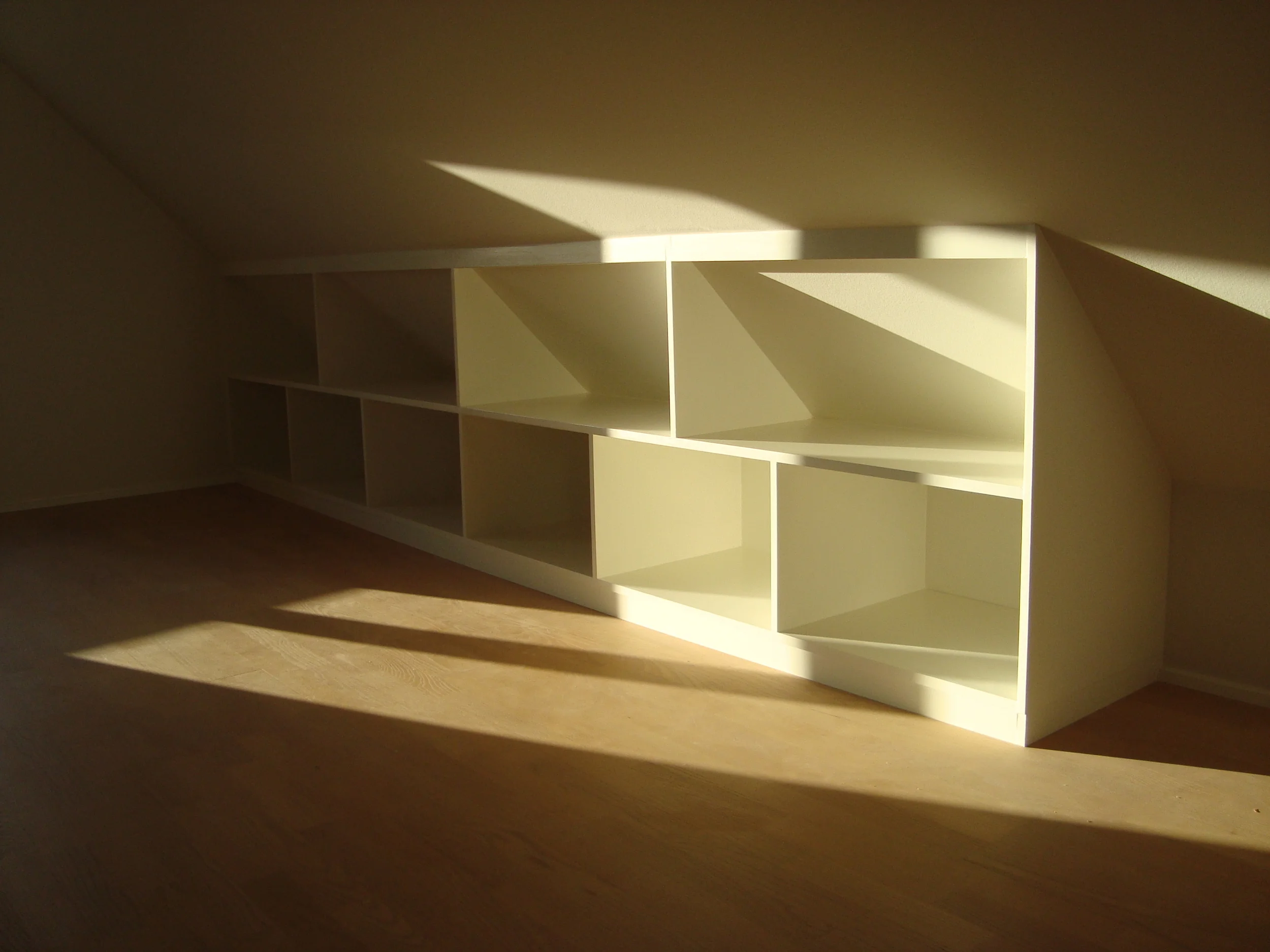 Hvidt reolsystem med otte åbne rum, tilpasset en væg under et skråtag, med sollys der skinner ind og laver skygger på gulvet.