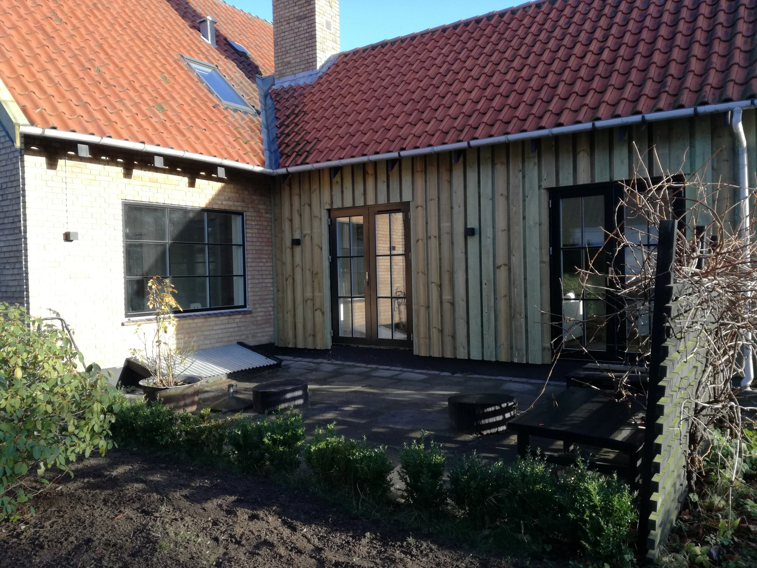 Nybygget terrasse med træmaterialer, døre og vinduer foran et hus med rødt tag og udendørs møbler.