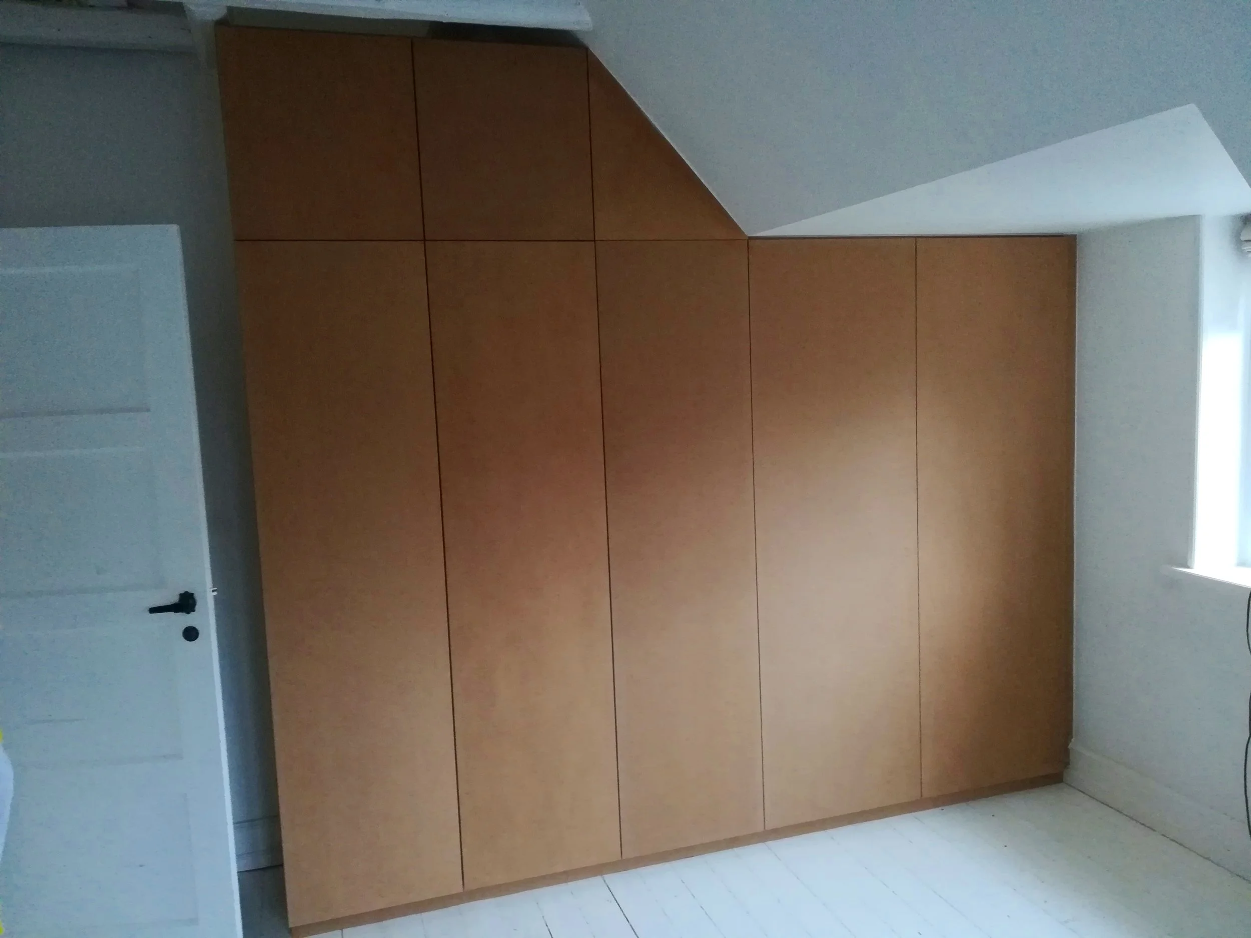 Et værelse med en stor garderobeskab med seks skabslåger, hvid væg og et vindue med gardin.