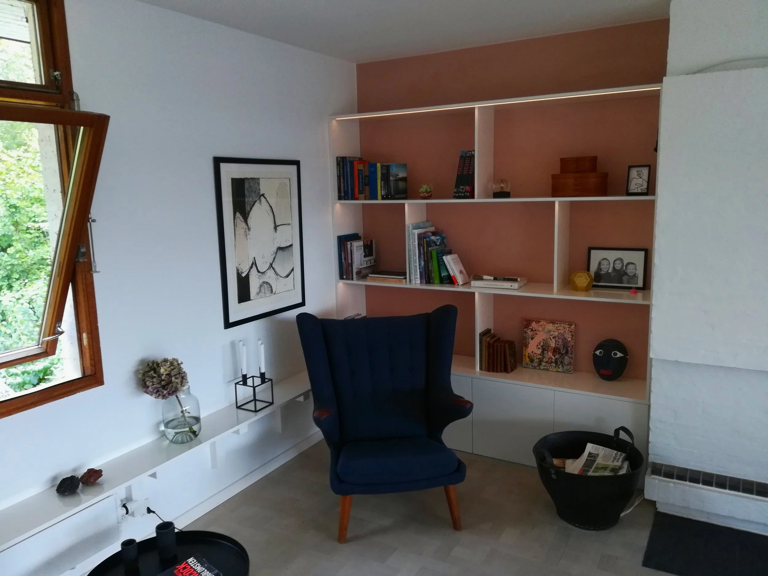 Et hjørne af et værelse med en åben vindue, en væg med et sort-hvidt abstrakt billede, en hvid bogreol med bøger og dekorative genstande, et blåt armstol, en sort kurv med aviser, og en hvid skænk med blomster og stearinlys.