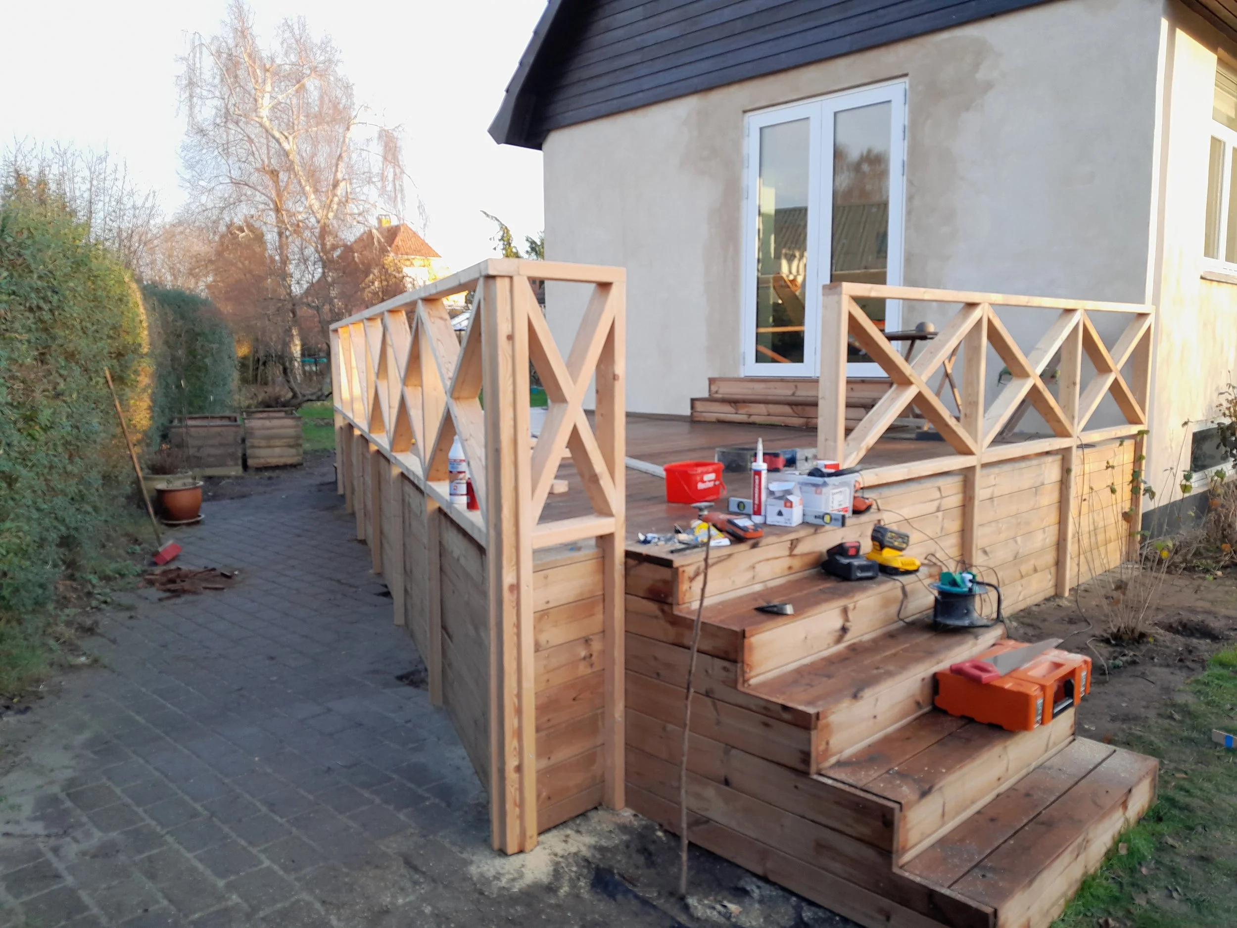 Udendørs træterrasse under opbygning udenfor et hus, med værktøj og materialer, og nye trappetrin, mens arbejdsprocessen er i gang.