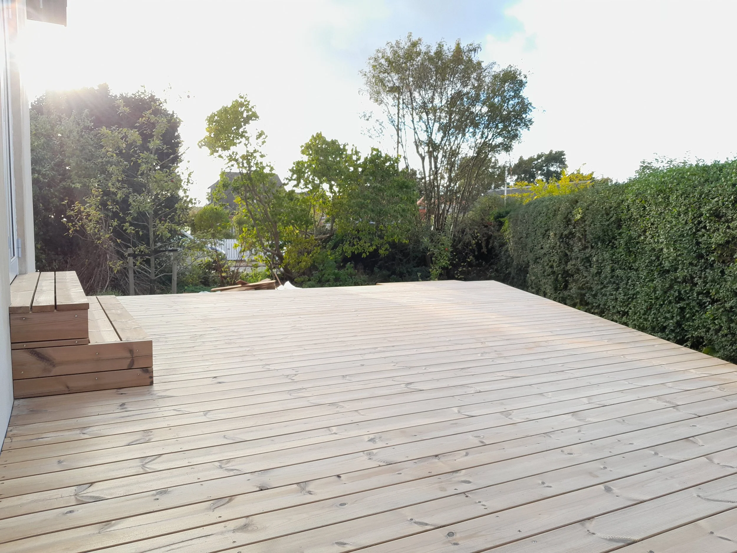 Nyt trædæk udendørs med grønne buske og træer i baggrunden, solskin og en hvid bygning til venstre.