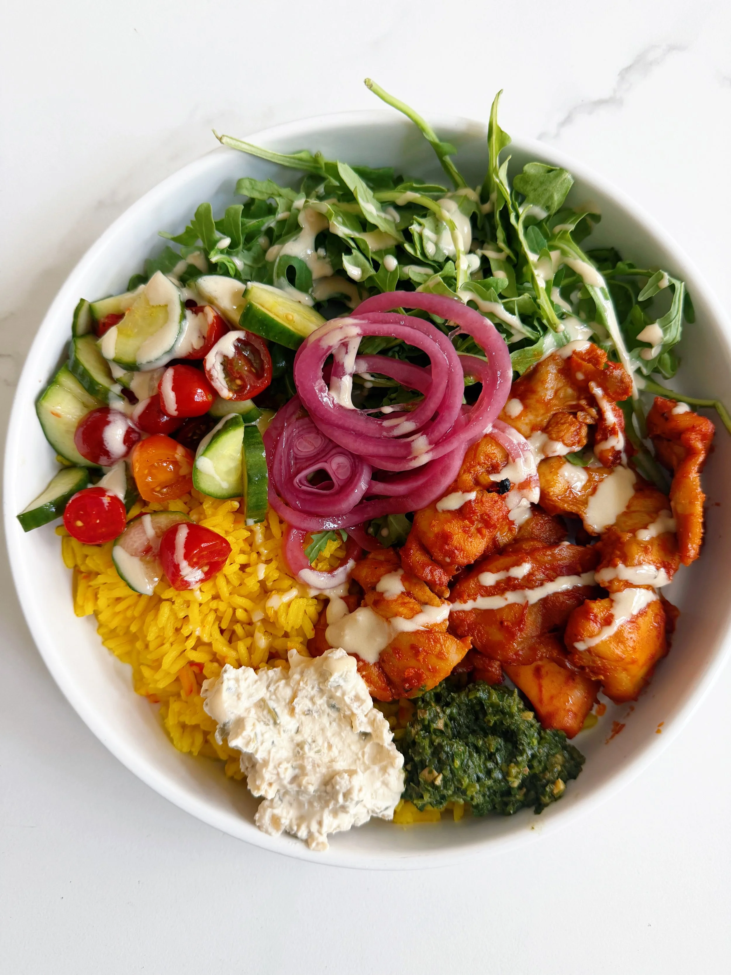 bowl filled with harissa chicken, arugula, cucumber tomato salad, and various sauces