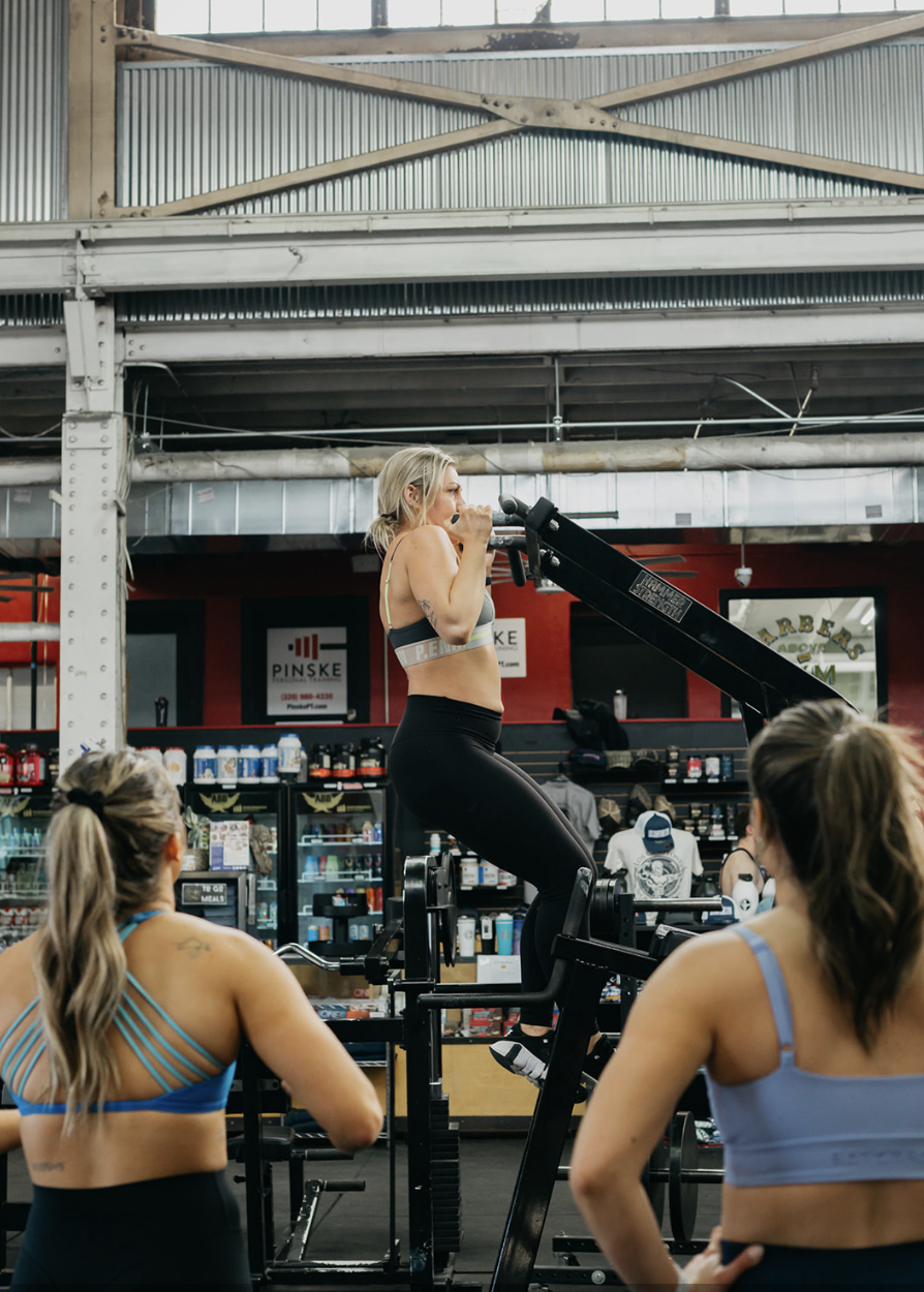 A woman in a sports bra and leggings using a rowing machine in a gym, with two women in sports bras observing her.