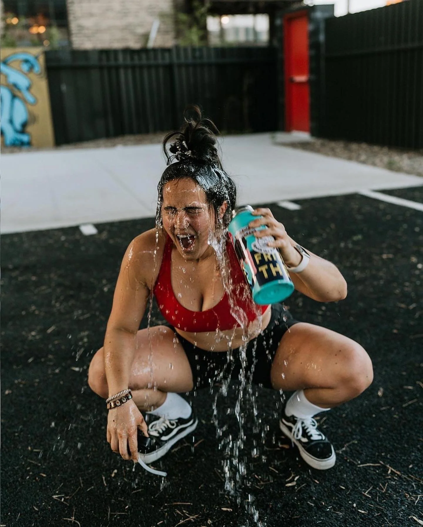 A young woman in a red sports bra, black shorts, and black and white sneakers is squatting on a black pavement, pouring water from a can and making a splash. She appears to be playing and having fun outdoors in a recreational area.