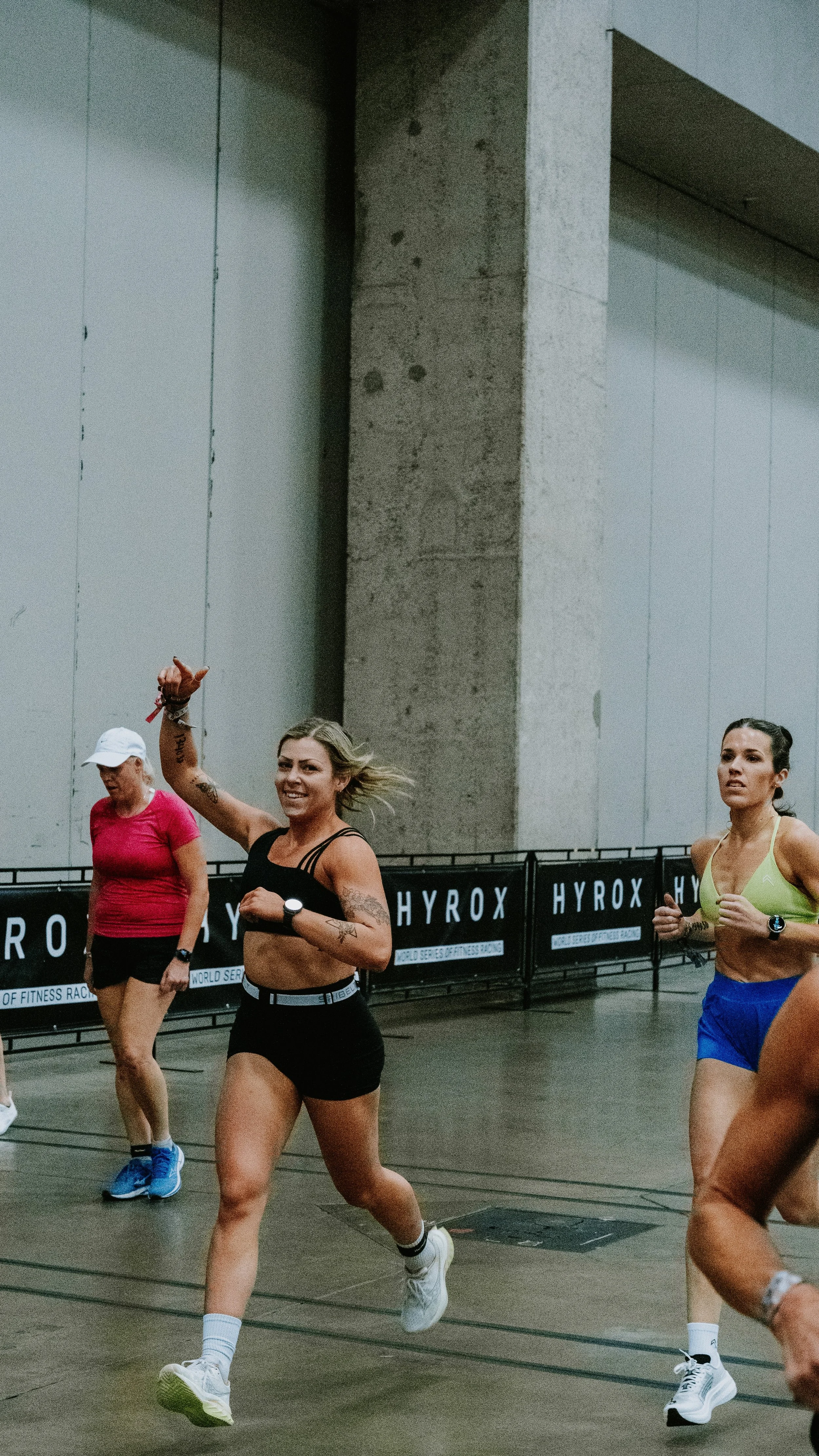 Women participating in a running race indoors, with one woman smiling and waving at the camera, wearing black athletic wear, while two others run nearby, one in pink and the other in yellow and blue shorts.