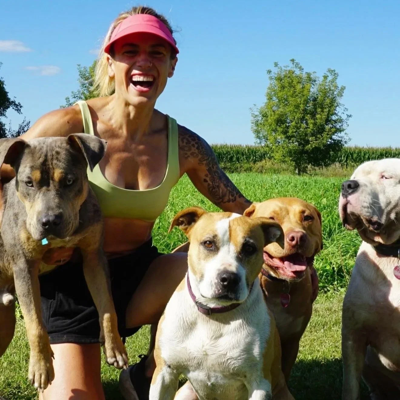 A woman with long blonde hair wearing a pink visor, beige tank top, and black shorts, smiling and surrounded by five dogs, outdoors on a sunny day with green grass, trees, and a blue sky.