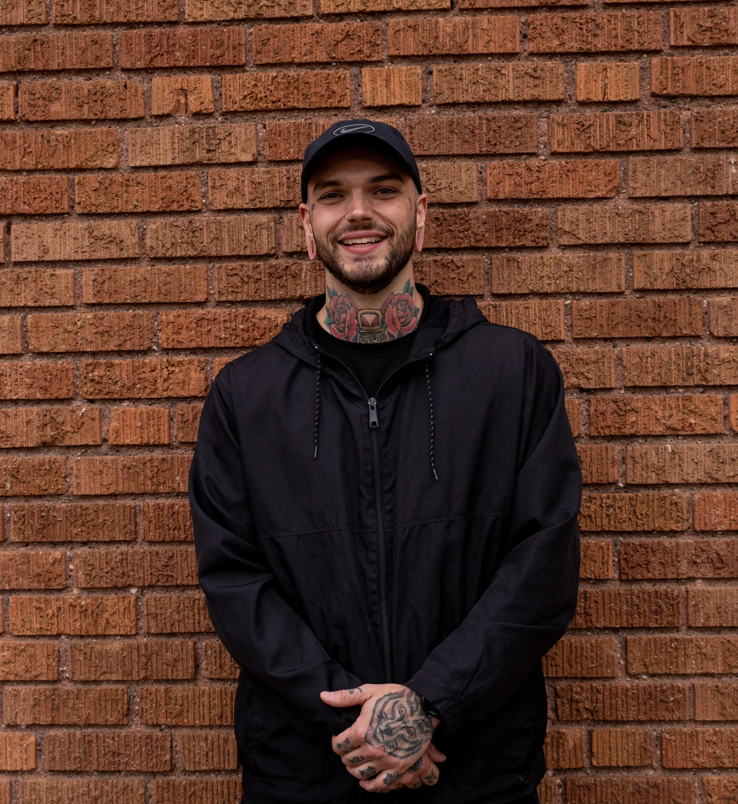 A smiling man standing against a brick wall, wearing a black jacket and hat, with tattoos on his neck and hands.