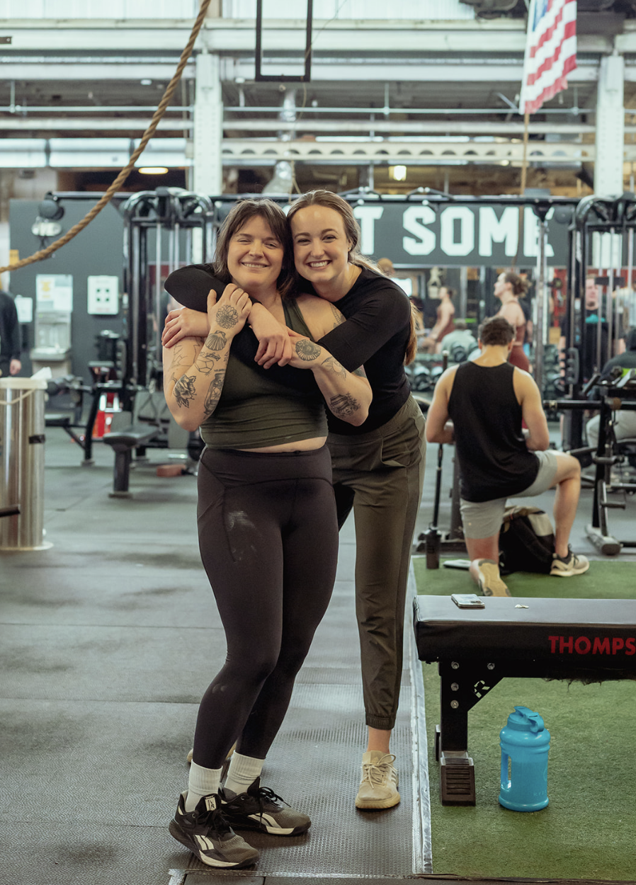 Two women hugging and smiling inside a gym with workout equipment and people exercising in the background.
