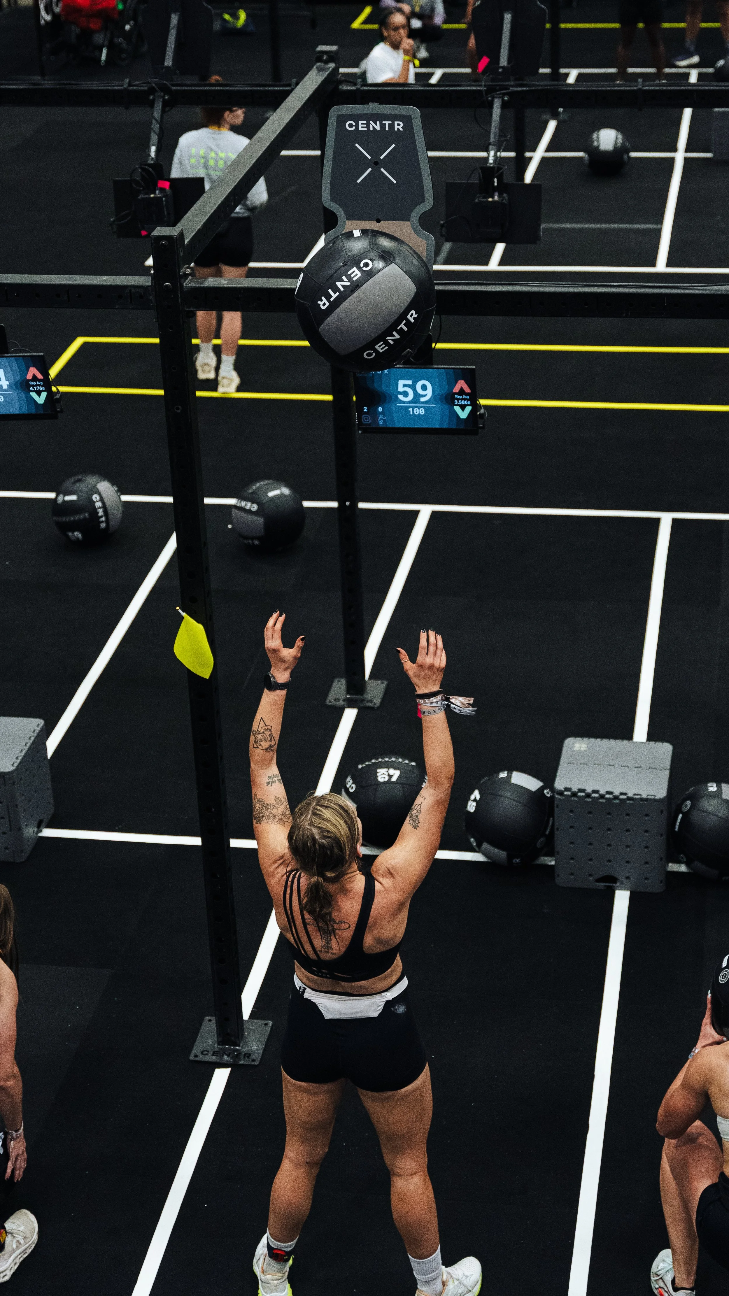 A woman with tattoos and a sports watch is in a gym, preparing to do a workout with a ball hanging from a high bar, with others in the background and a digital screen displaying the number 59.