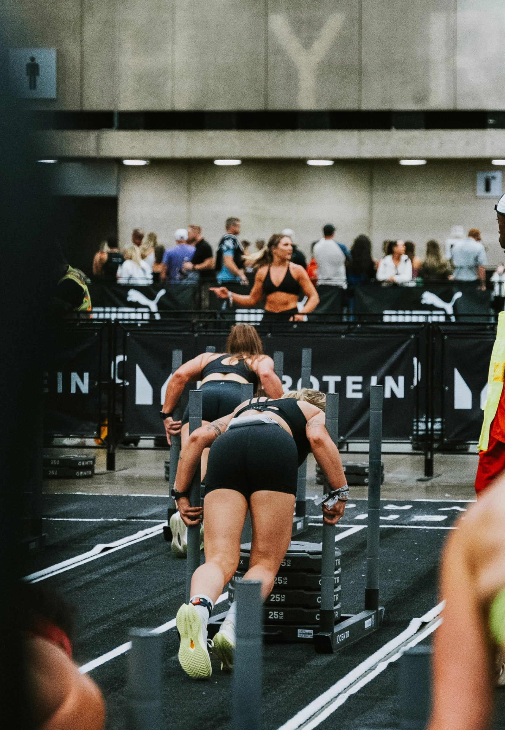 Two women performing push-ups on push-up bars at a fitness event with an audience watching in the background.