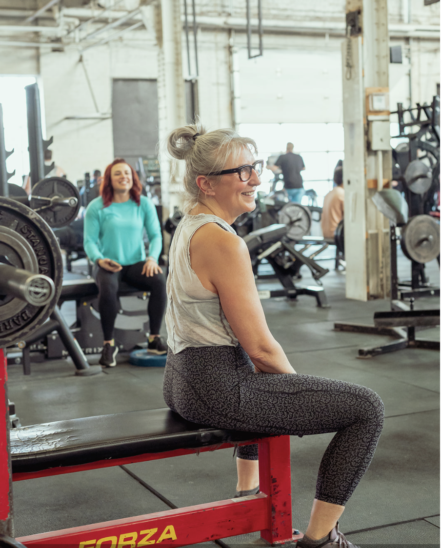 Two women at a gym, one sitting on a bench and smiling, the other sitting on a box, with various gym equipment around them.