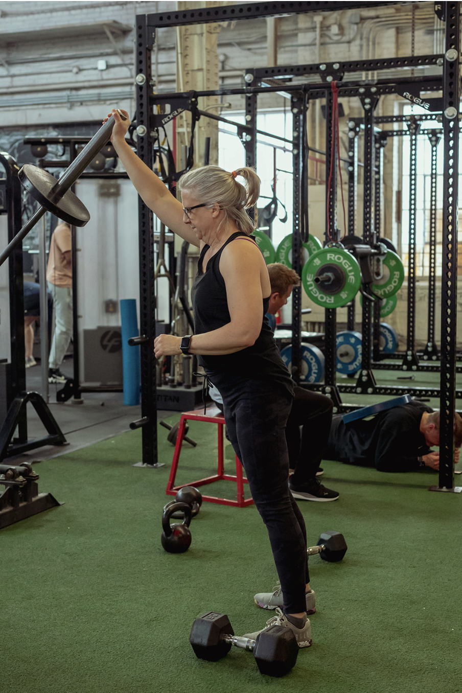A woman with gray hair in a ponytail, wearing glasses and a black sports tank top, is lifting a dumbbell overhead at a gym. She is standing on a green turf floor surrounded by gym equipment, including kettlebells on the ground and weight plates on a rack in the background.