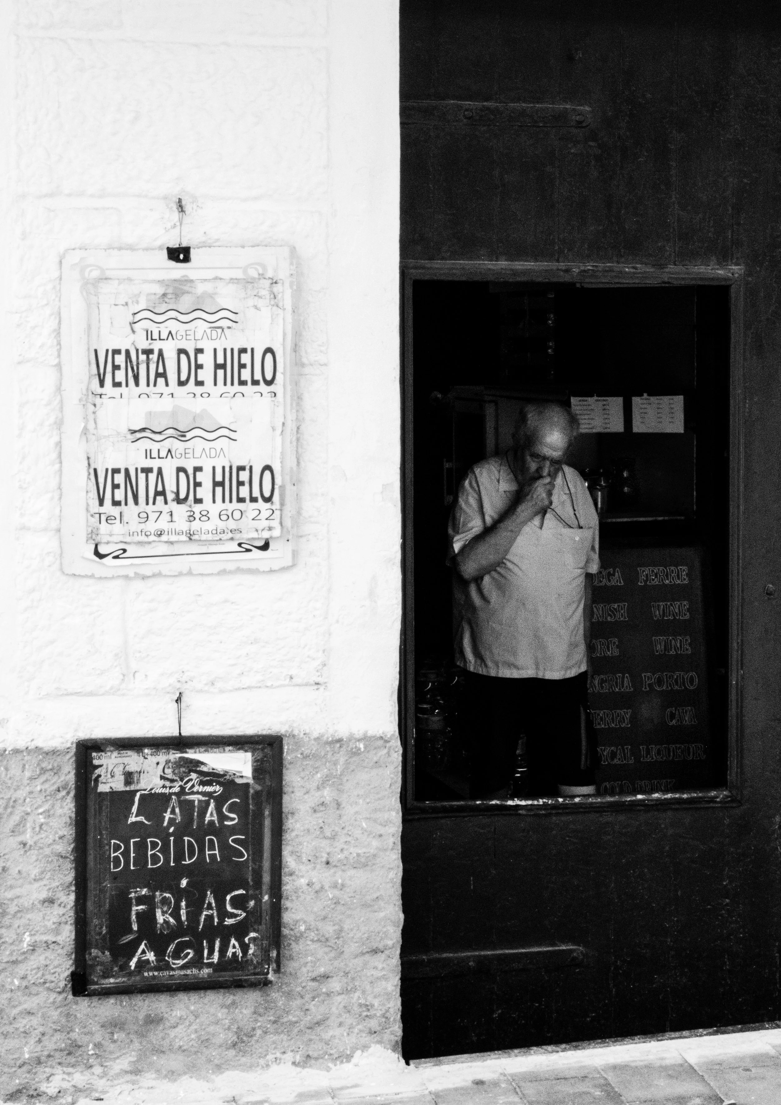 Street scene showing a man inside a shop behind the window, with various items on shelves. Outside, signs advertise ice cream sales and drinks in Spanish. The wall is textured with peeling paint, and the area looks aged and rustic.