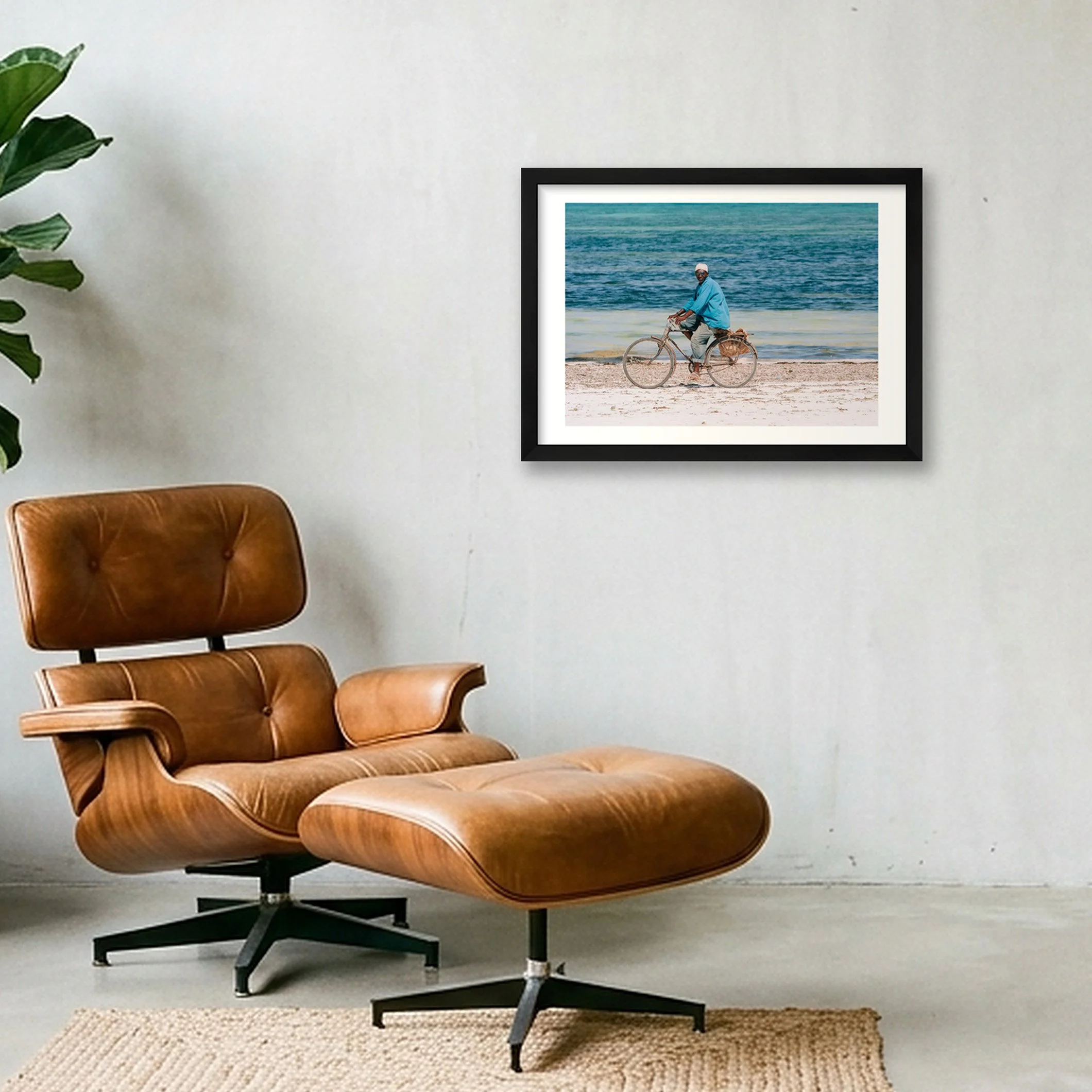 A leather lounge chair near a green plant and a framed photograph of a person riding a bicycle on a beach hangs on a plain white wall, with a woven rug on the floor.