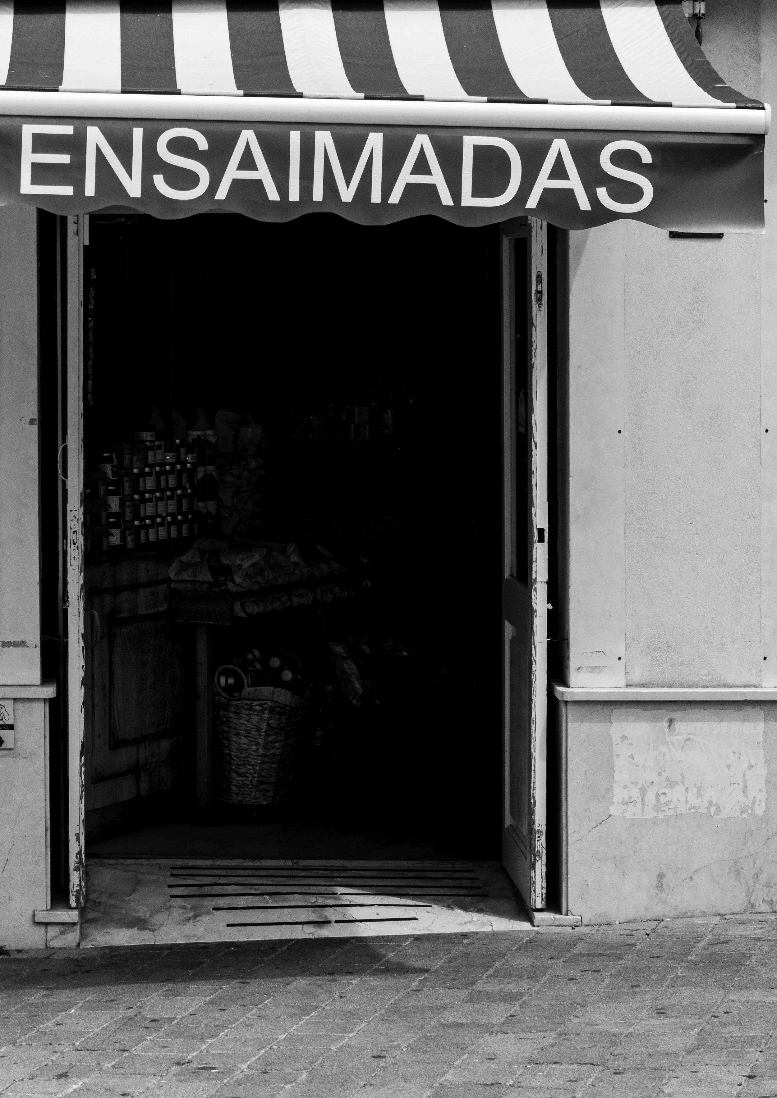A storefront with a striped awning and a sign that reads 'ENSAIMADAS'; the store appears to be a small shop selling food or baked goods, with some items visible inside.