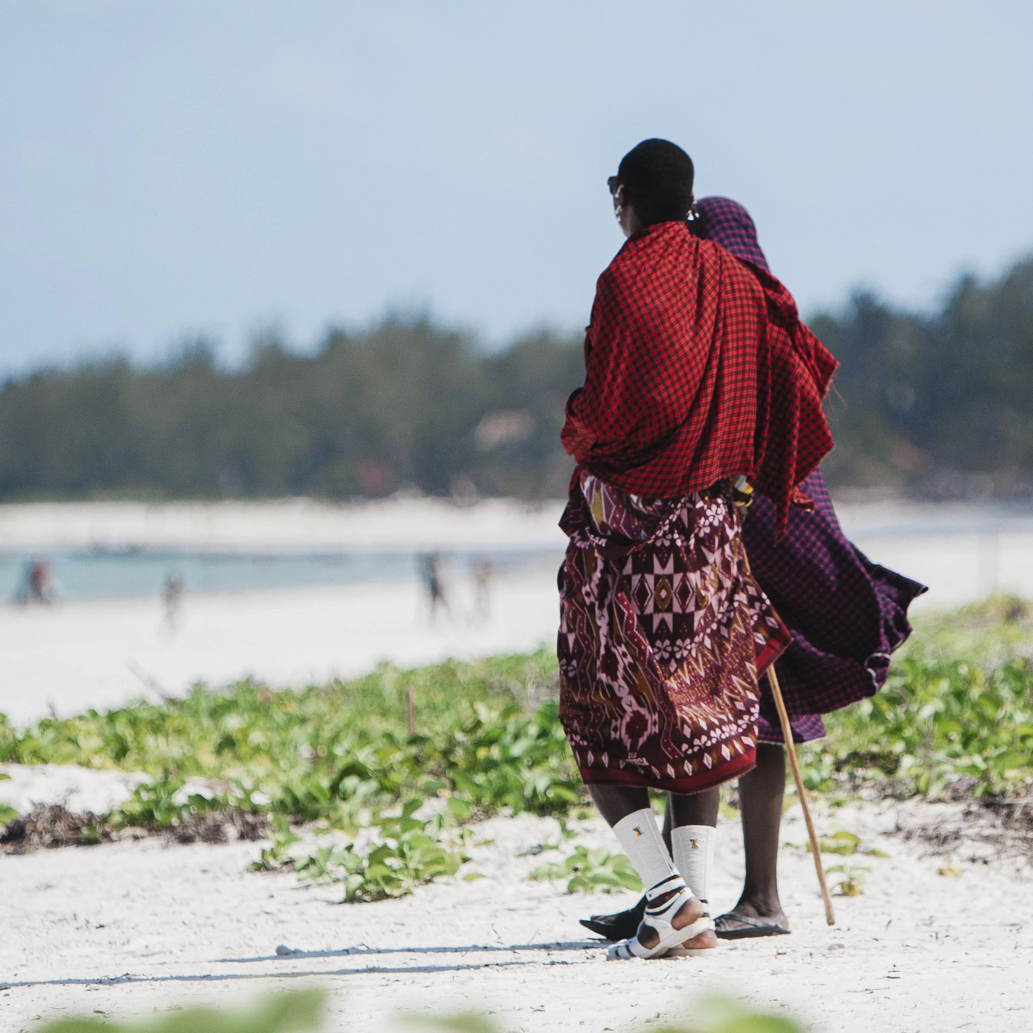 Two Maasai women walking on a beach, dressed in traditional colorful shukas and sandals, with one using a walking stick.
