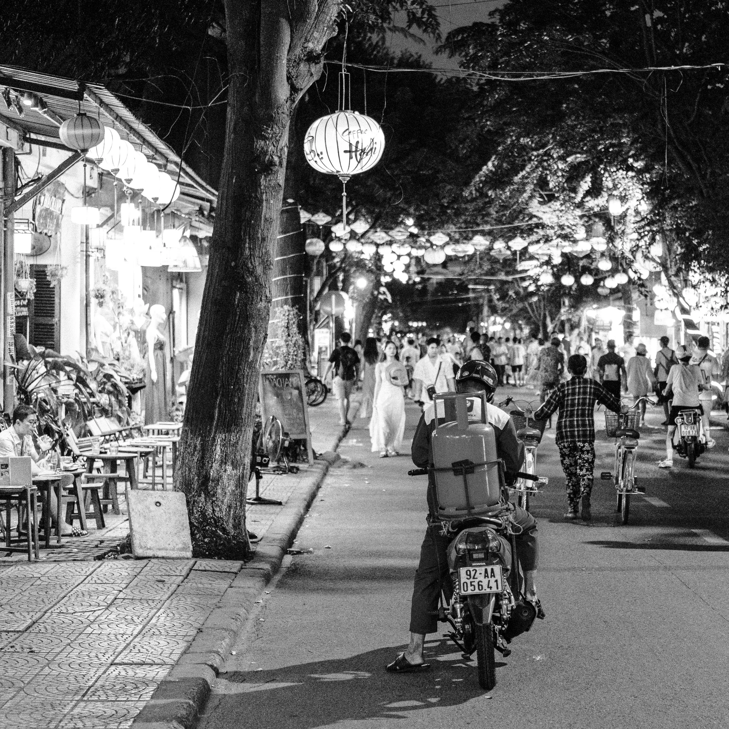 A nighttime street scene with lanterns hanging overhead, people walking and cycling, and outdoor seating along the sidewalk, in black and white.