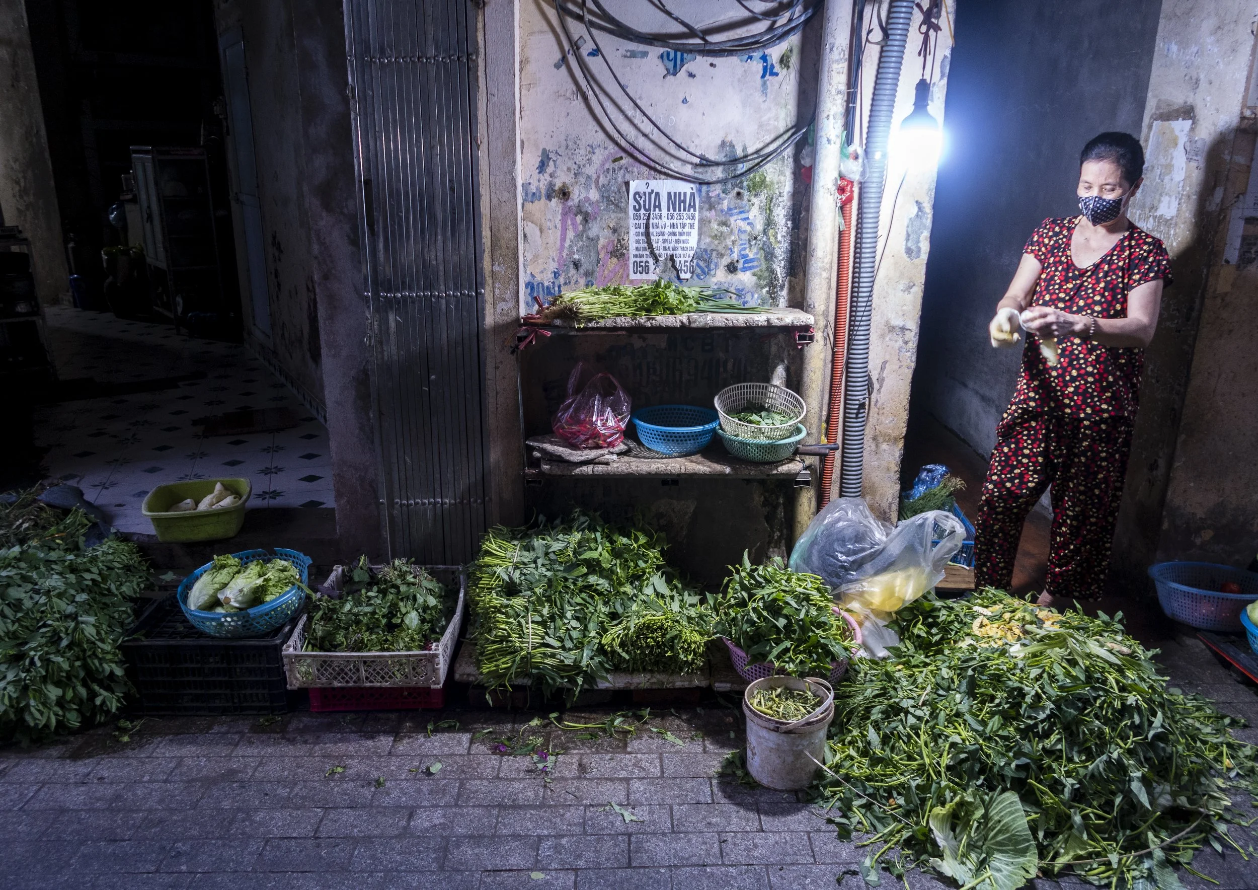 Street vendor selling fresh vegetables including leafy greens on a small outdoor stand. A woman wearing a mask and floral pajamas is preparing the vegetables. The stand is set against a weathered wall with a sign in Vietnamese. The ground is tiled and scattered with leaves and vegetable scraps.