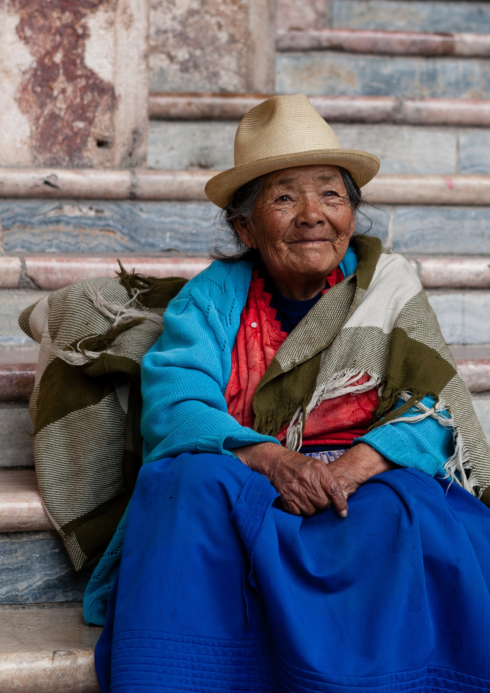 An elderly woman sitting on stone stairs, wearing a beige hat, a multicolored shawl, a bright blue sweater, and blue pants, smiling gently at the camera, with a weathered stone wall in the background.