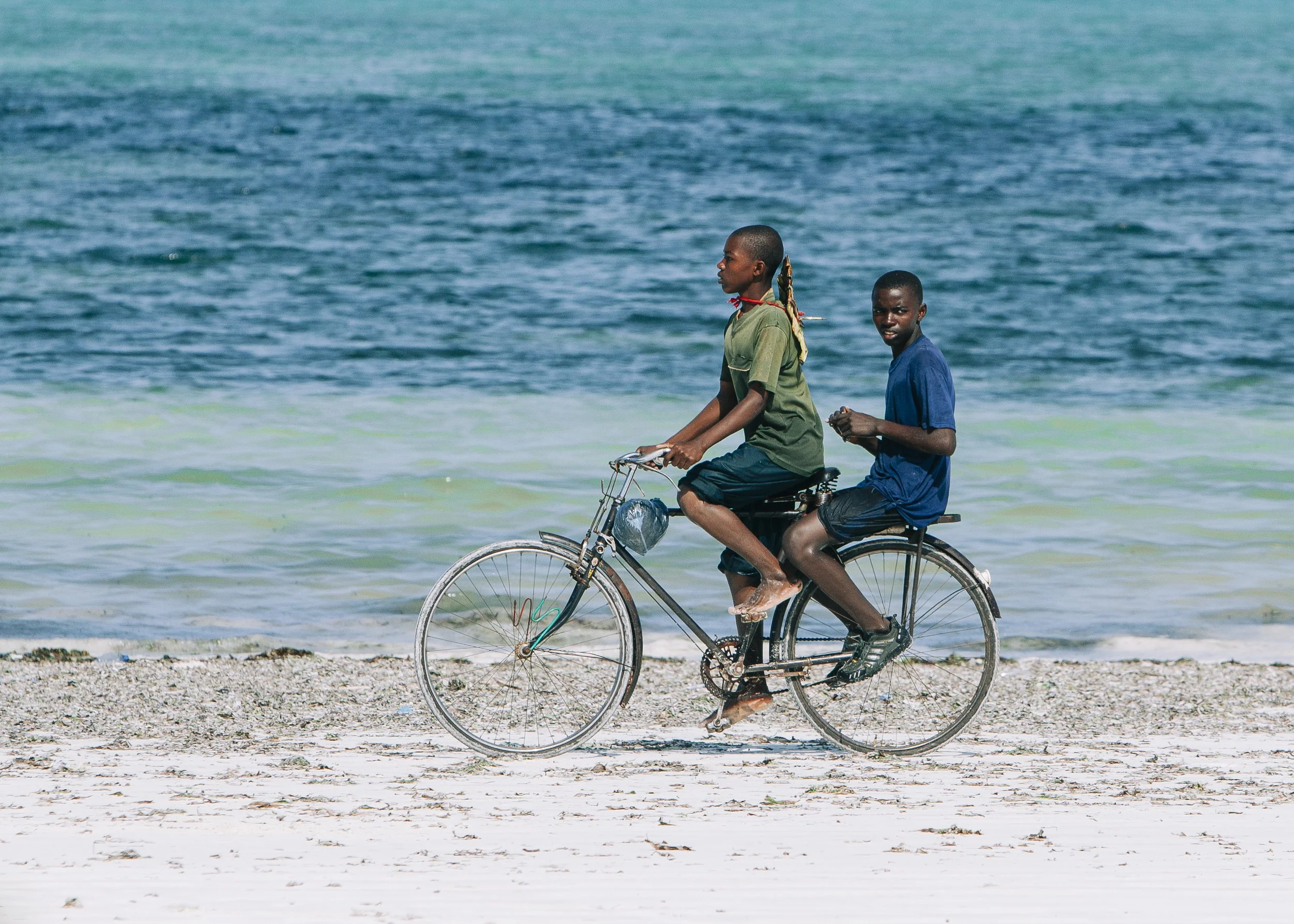 Two boys riding a bicycle along a beach with the ocean in the background. One boy pedals, while the other sits on the rear of the bike.