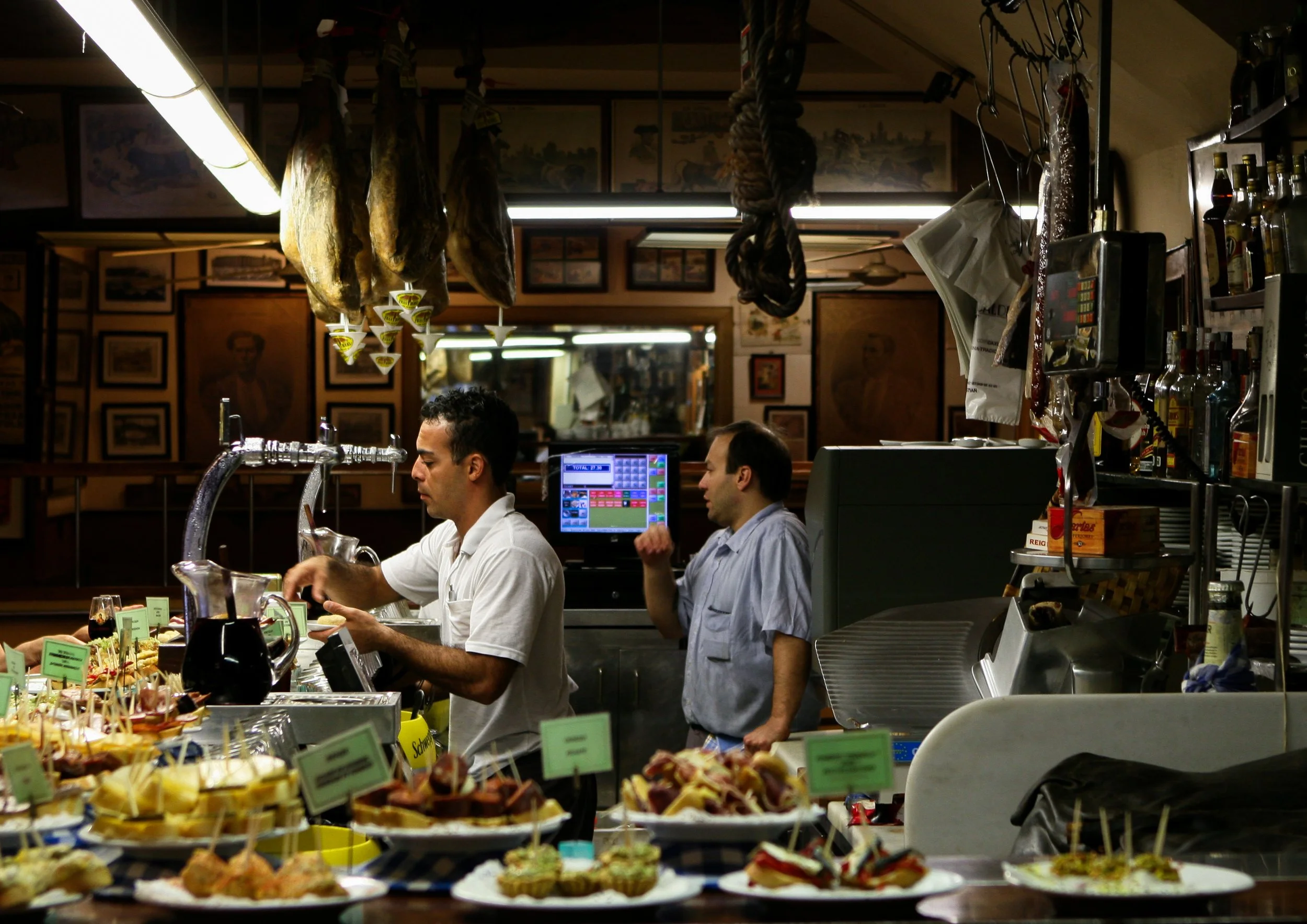 Interior of a deli or bakery with two men working behind a counter, surrounded by plates of food and various supplies.