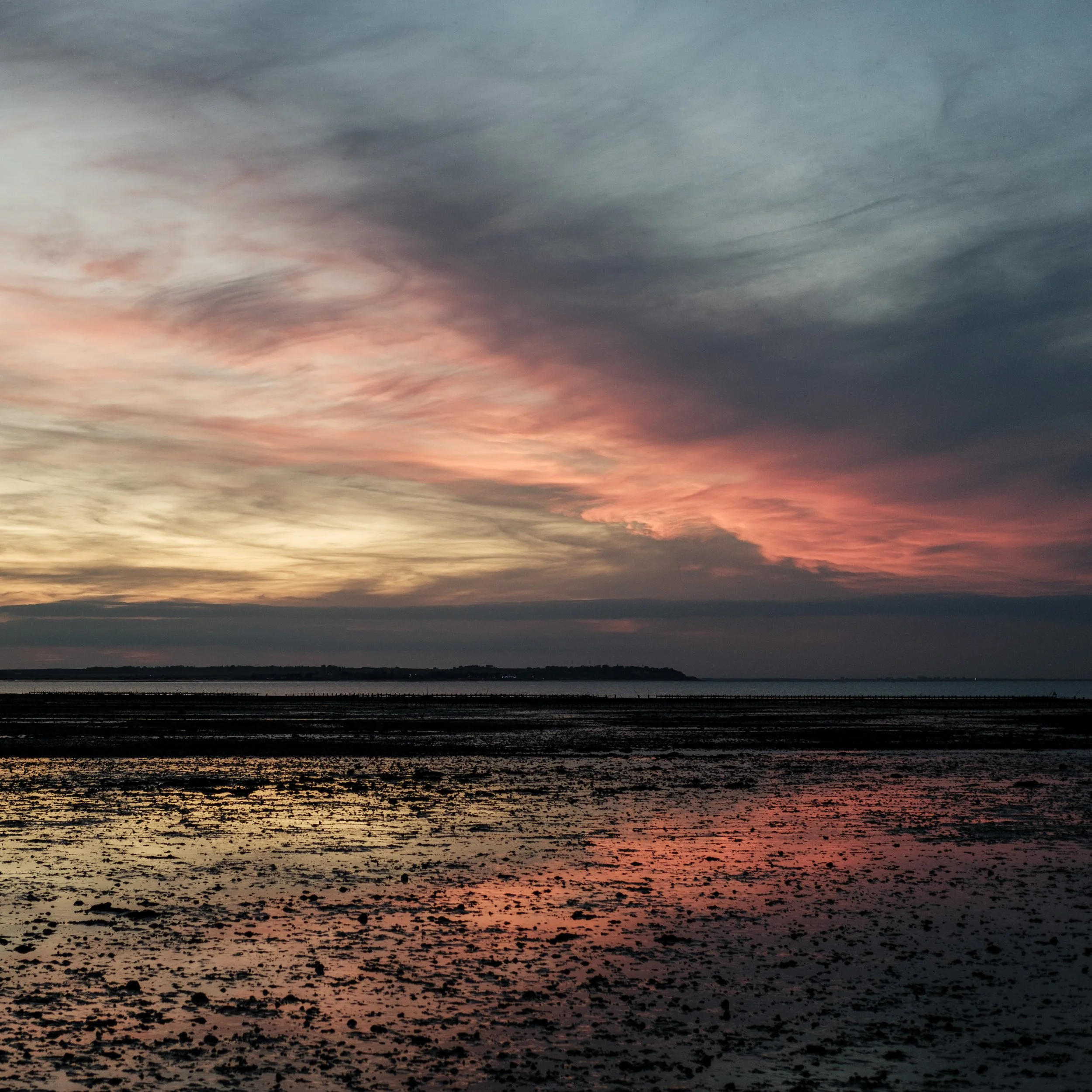 Dramatic pink and orange sunset over a calm coastal tidal flat in whitstable with silhouetted shoreline and reflective wet sand