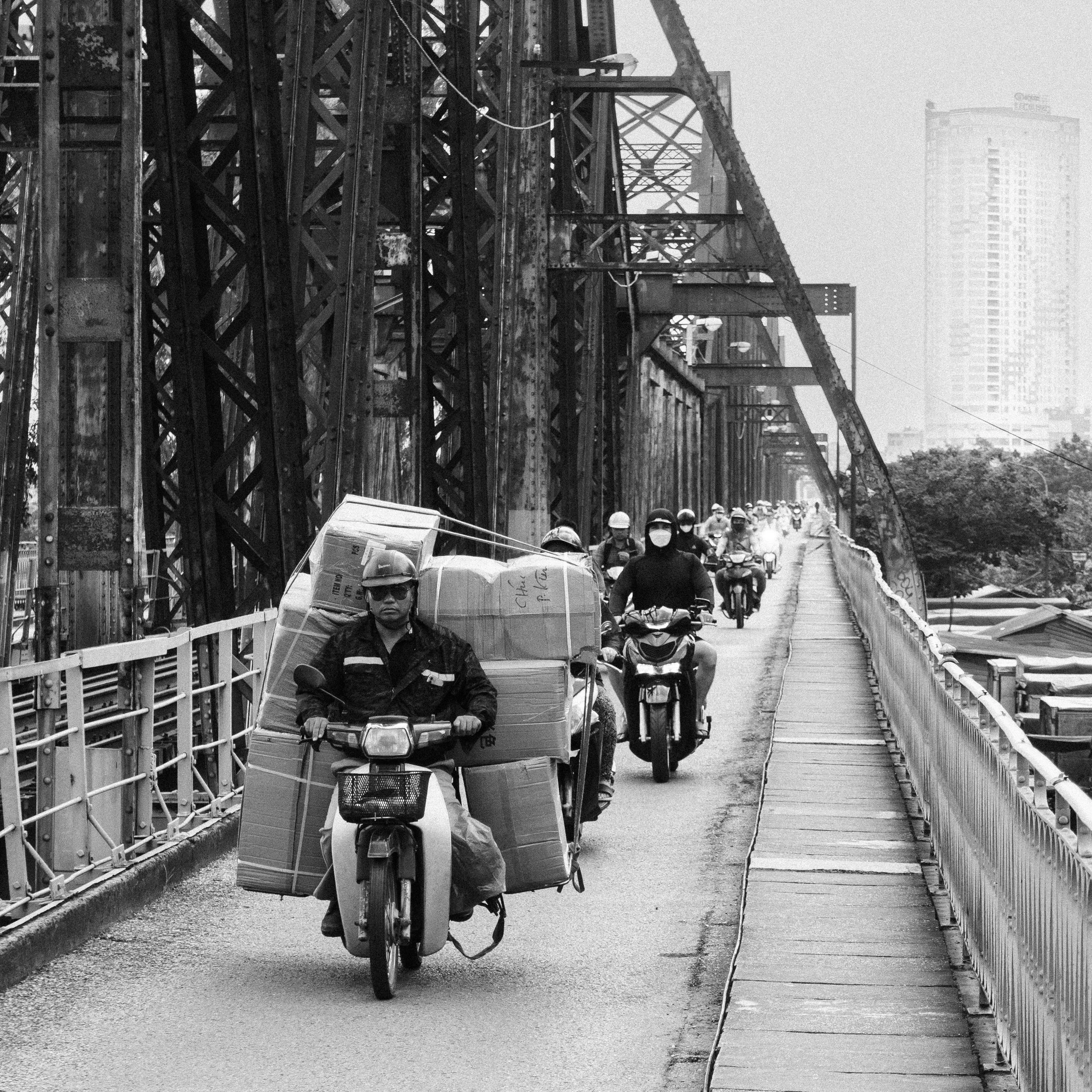 A group of motorcyclists riding across a bridge with a steel structure, some wearing helmets and face masks, in an urban area with tall buildings in the background.