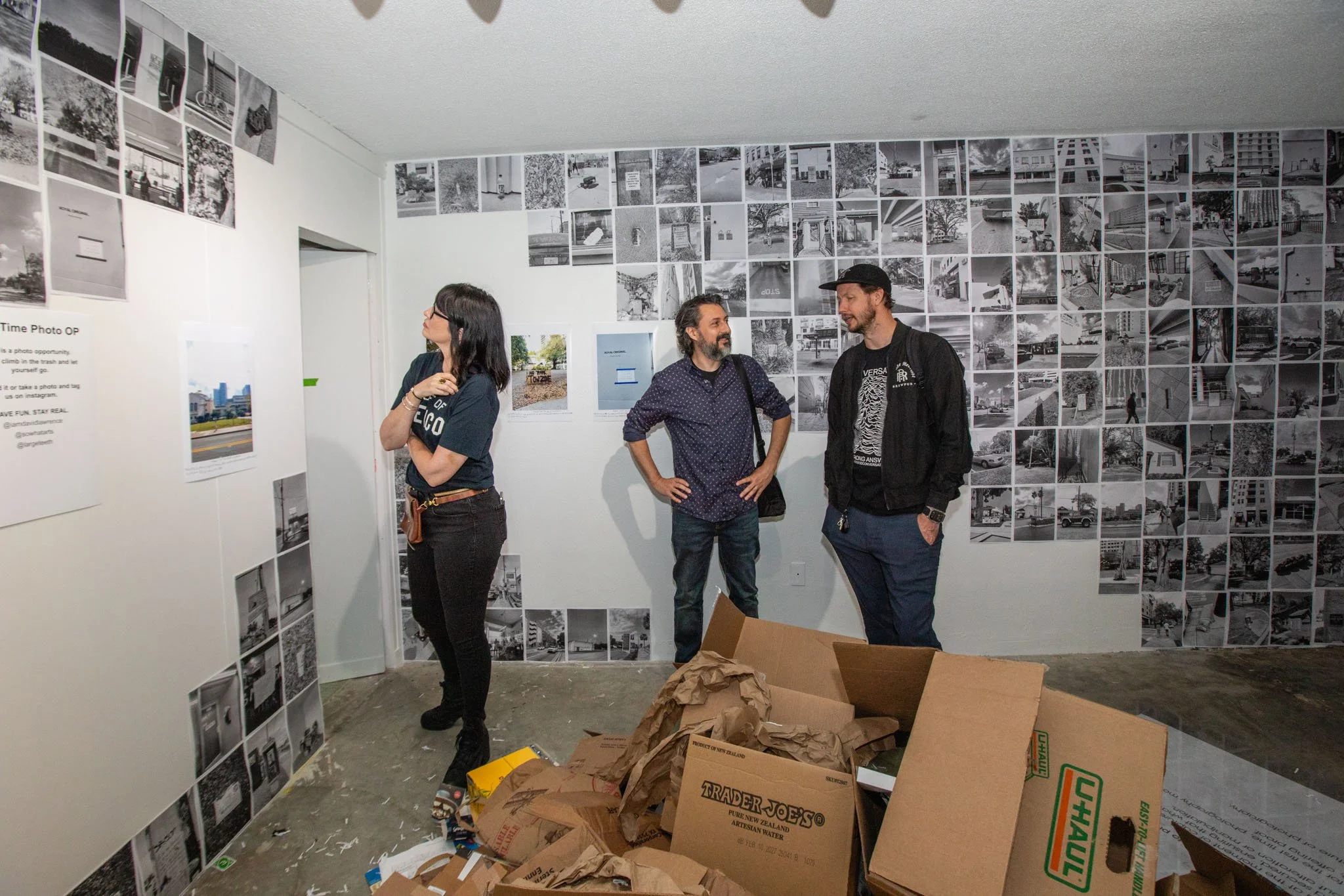 Three people standing inside an art gallery or exhibition space looking at black and white photographs on the wall. The wall features a collage of multiple photographs, and there are empty boxes and packing materials on the floor.