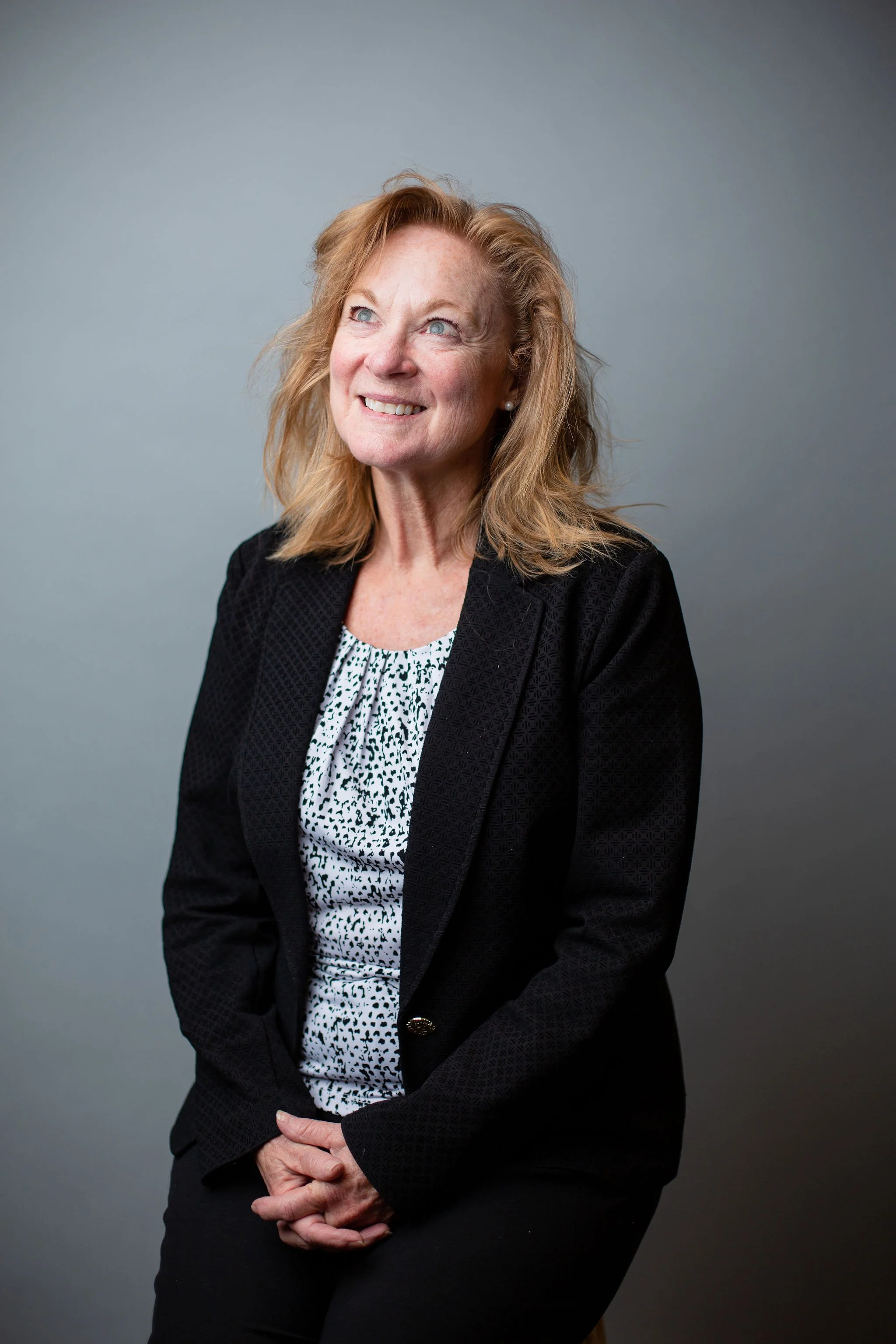A senior woman with red hair and blue eyes sitting against a gray background, smiling slightly, wearing a black blazer over a white and black patterned blouse.