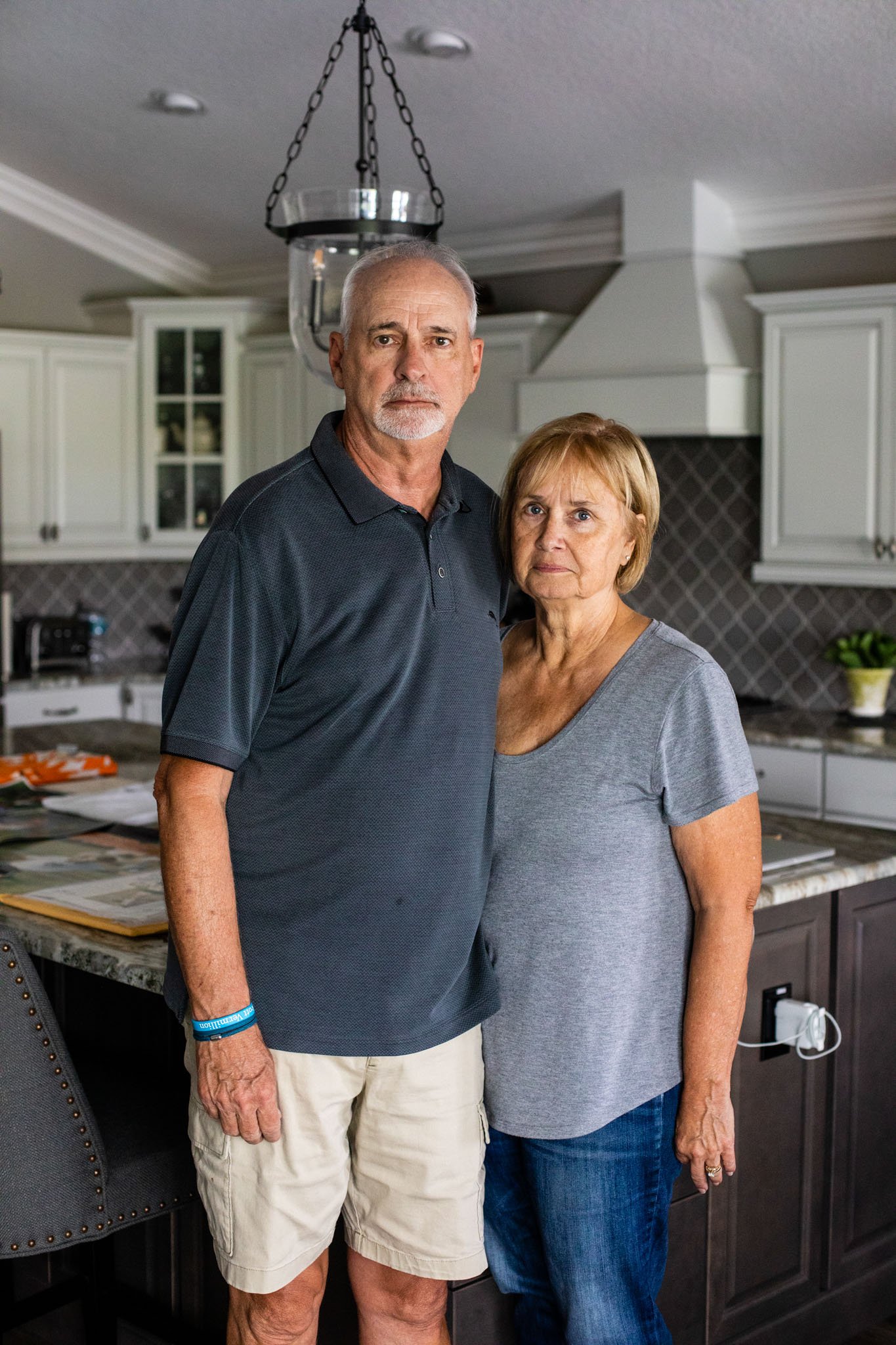 An elderly man and woman standing in a kitchen, looking at the camera with serious expressions.