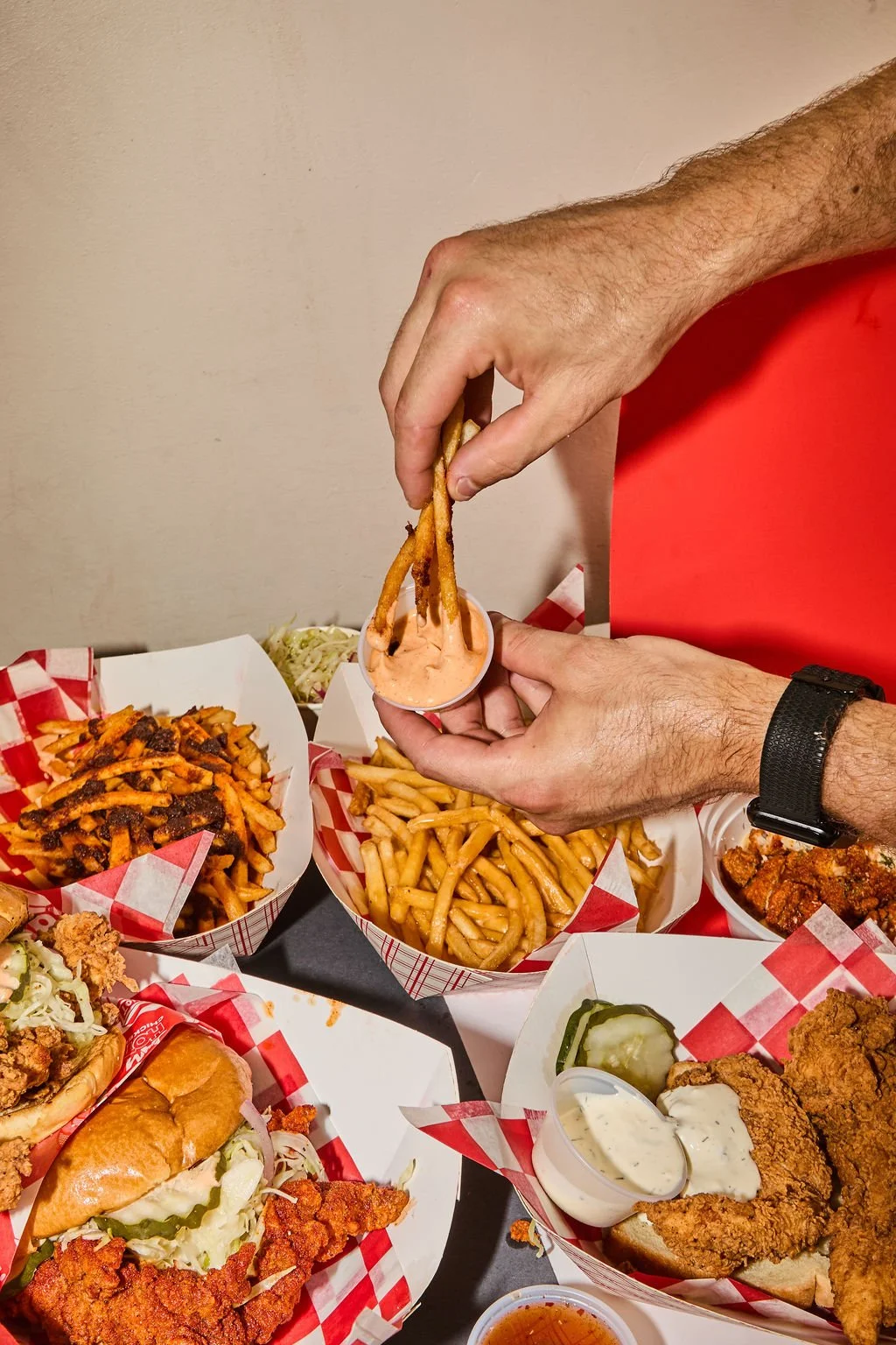 A person dipping French fries into a pink dipping sauce, surrounded by various fast food items like fries, fried chicken, a sandwich, and pickles on red and white checkered paper trays.