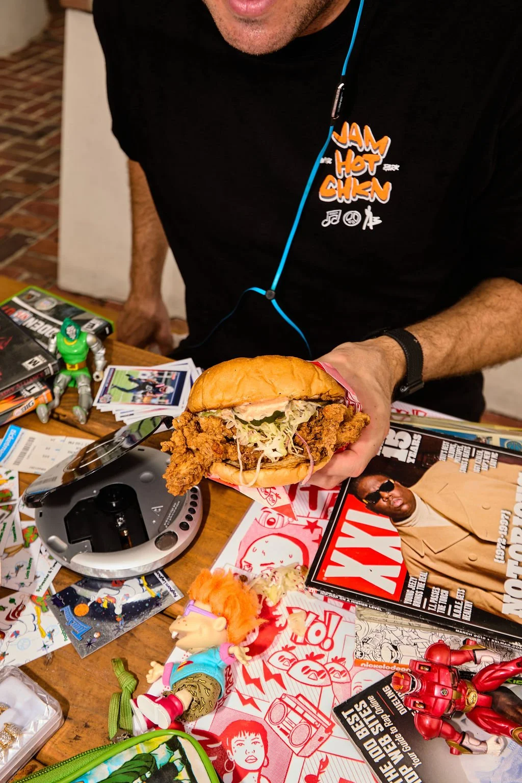 Person holding a fried chicken sandwich with lettuce and pickles. The table has toys, magazines, and various items.