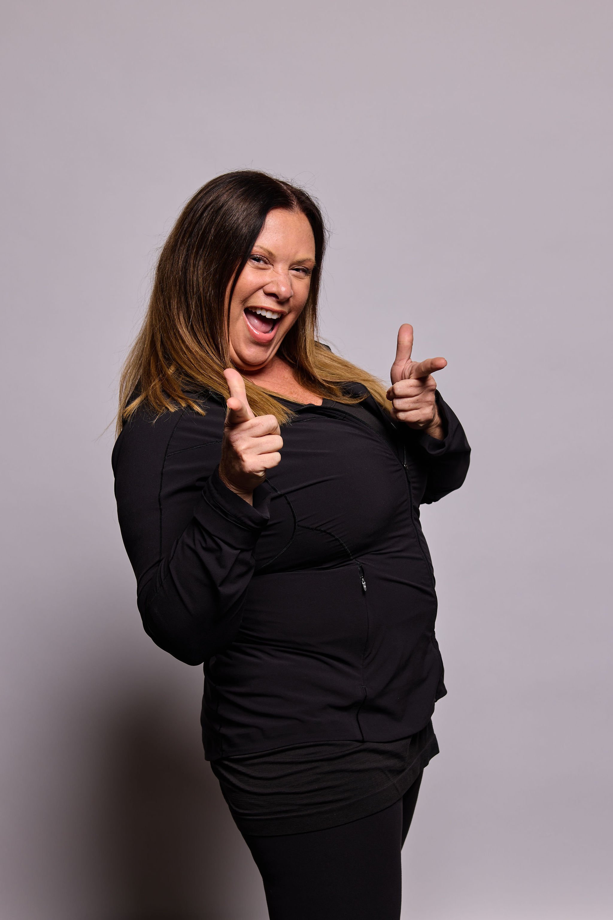 Joyful woman with long brown hair pointing both index fingers towards the camera, wearing a black jacket against a plain gray background.