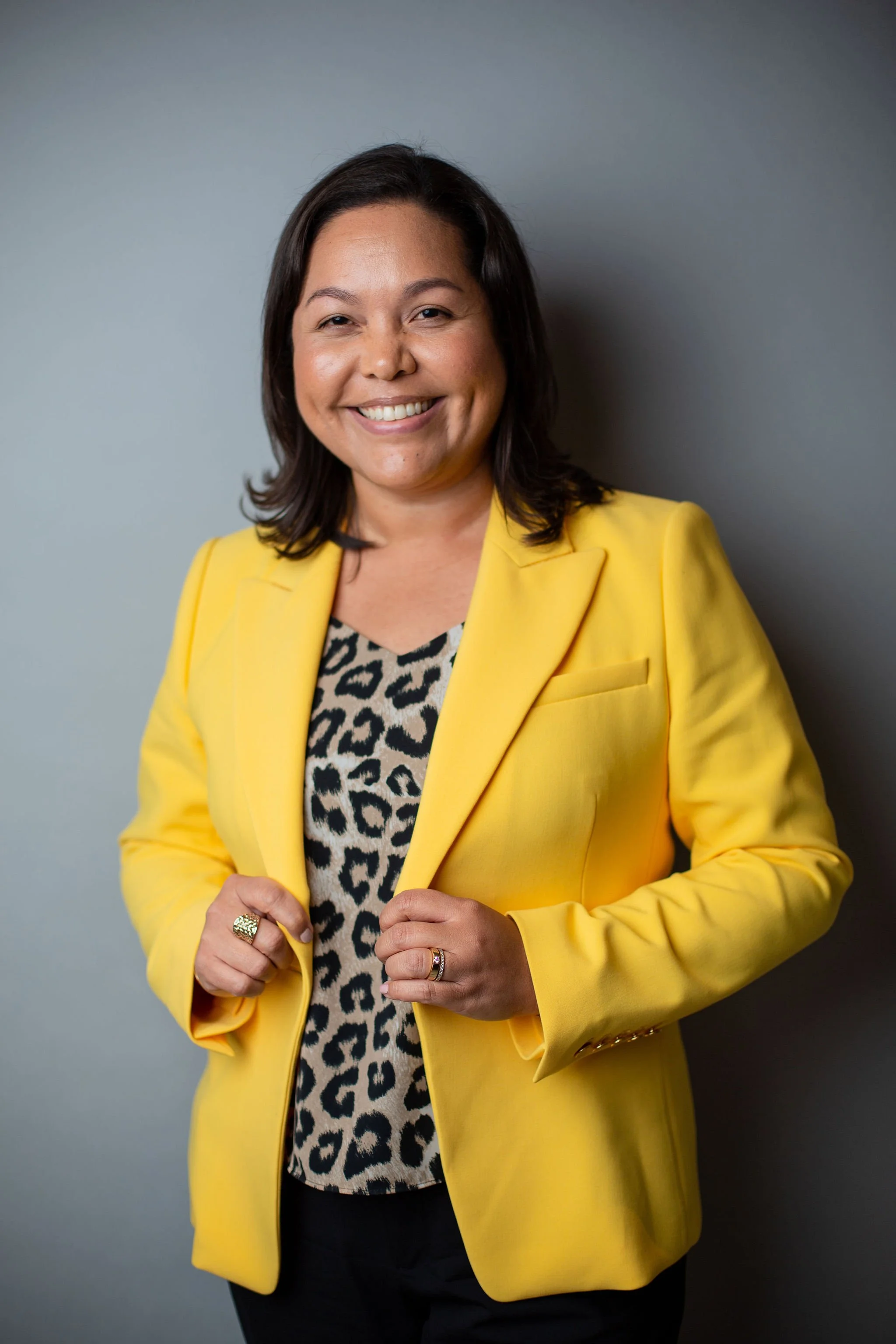 A smiling woman with shoulder-length dark hair wearing a yellow blazer over a leopard print top, against a plain gray background.