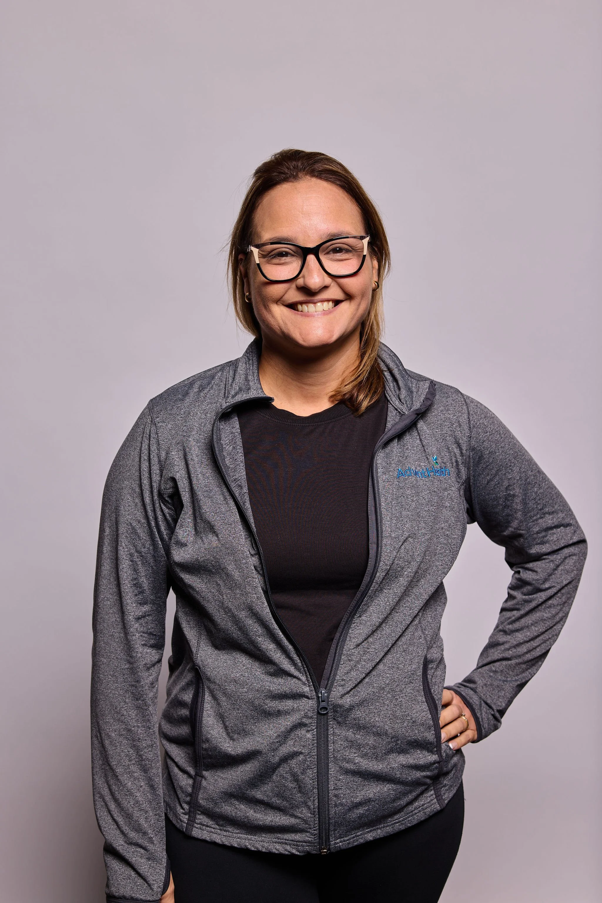 A smiling woman with glasses, wearing a gray zip-up athletic jacket over a black shirt, standing against a plain light gray background.