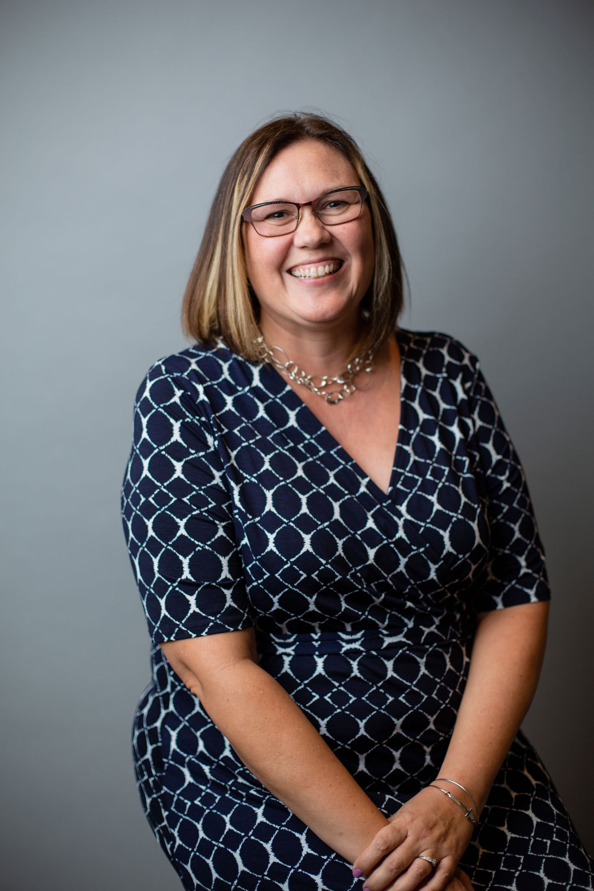 A woman with shoulder-length light brown hair, glasses, and a cheerful smile, wearing a navy blue dress with a white geometric pattern, sitting against a plain gray background.