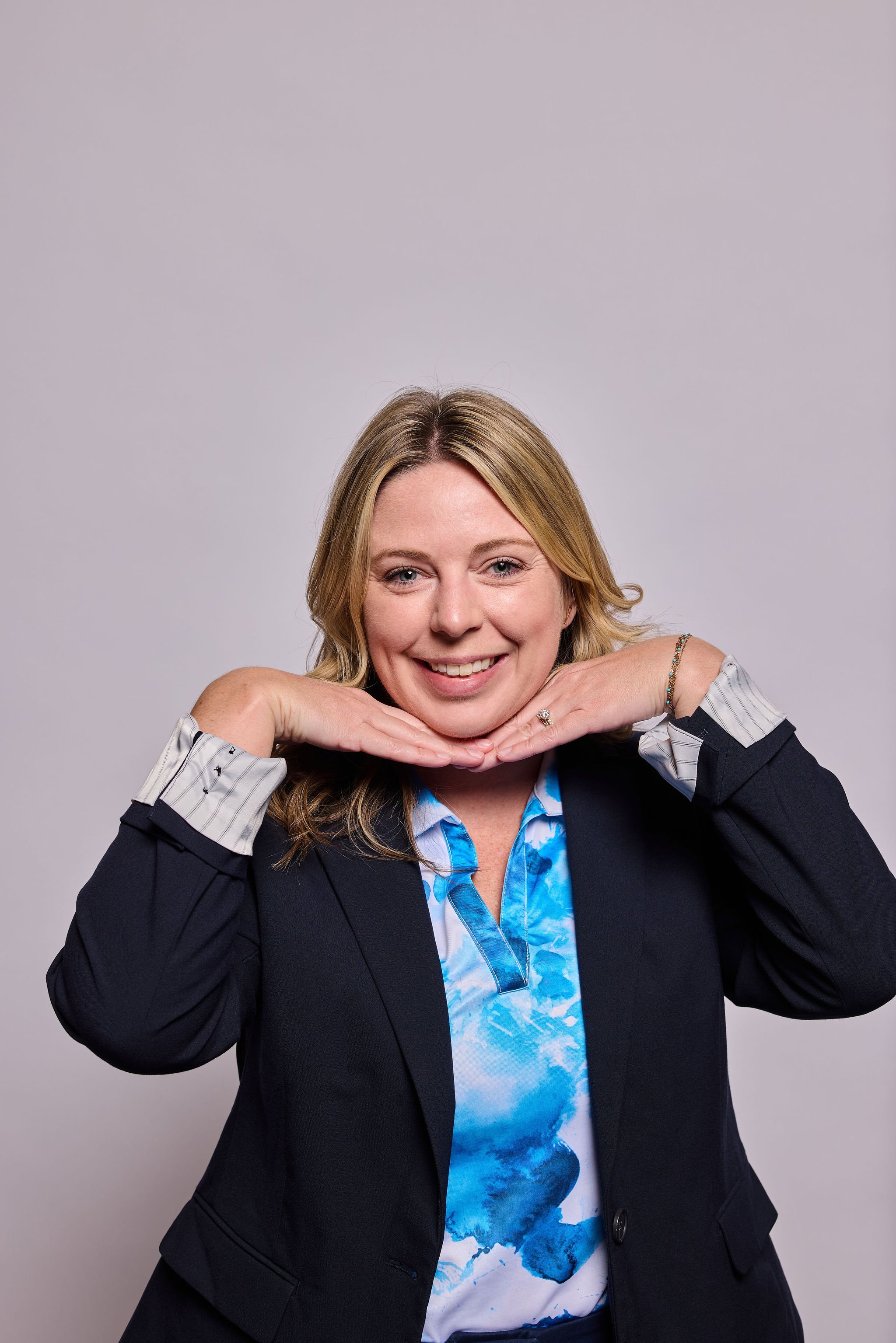 A woman with blonde hair smiling and resting her chin on her hands, wearing a black blazer over a blue and white floral shirt, against a light gray background.