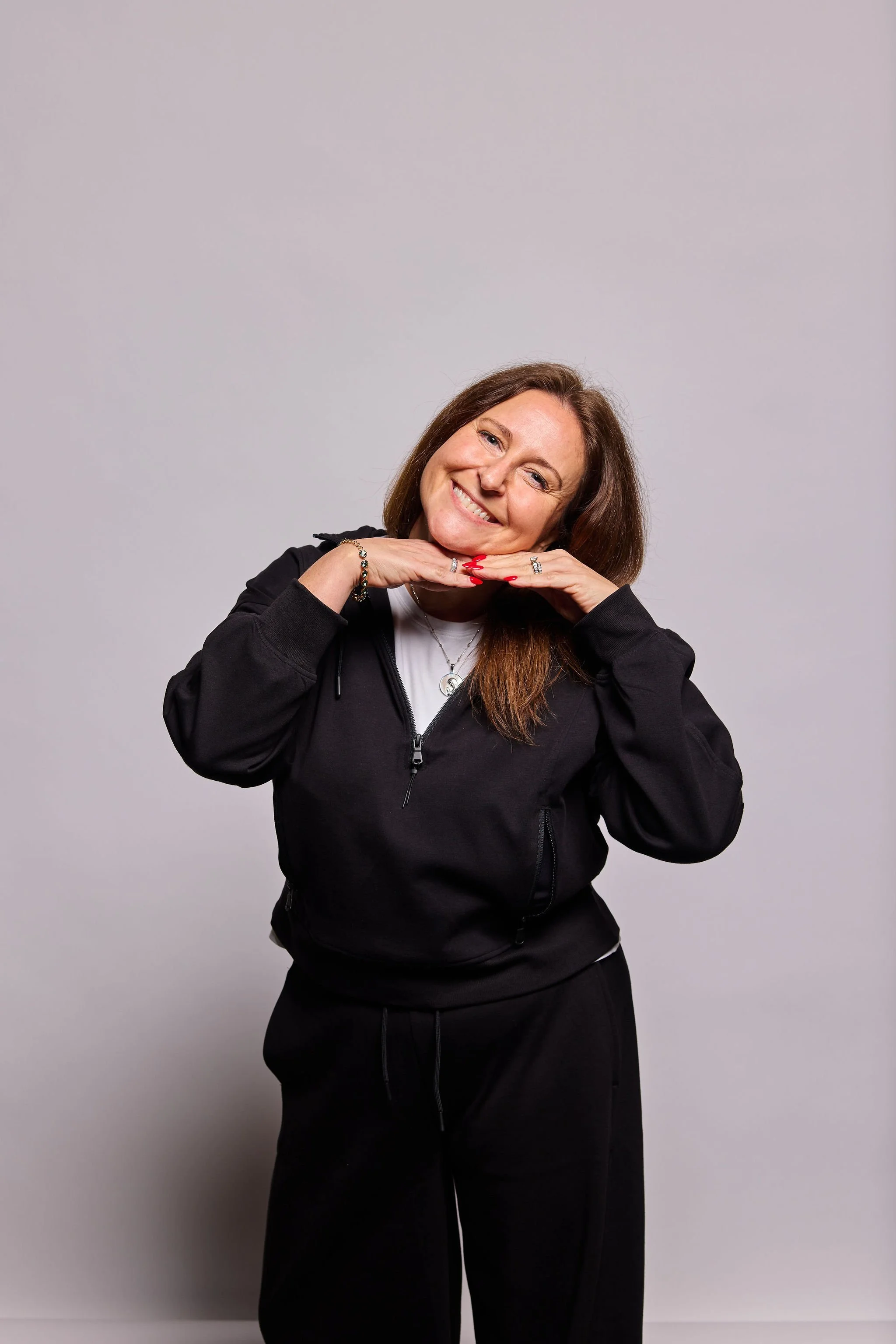 Smiling woman with brown hair posing with her chin resting on her hands, wearing a black hoodie and black pants, against a plain light gray background.