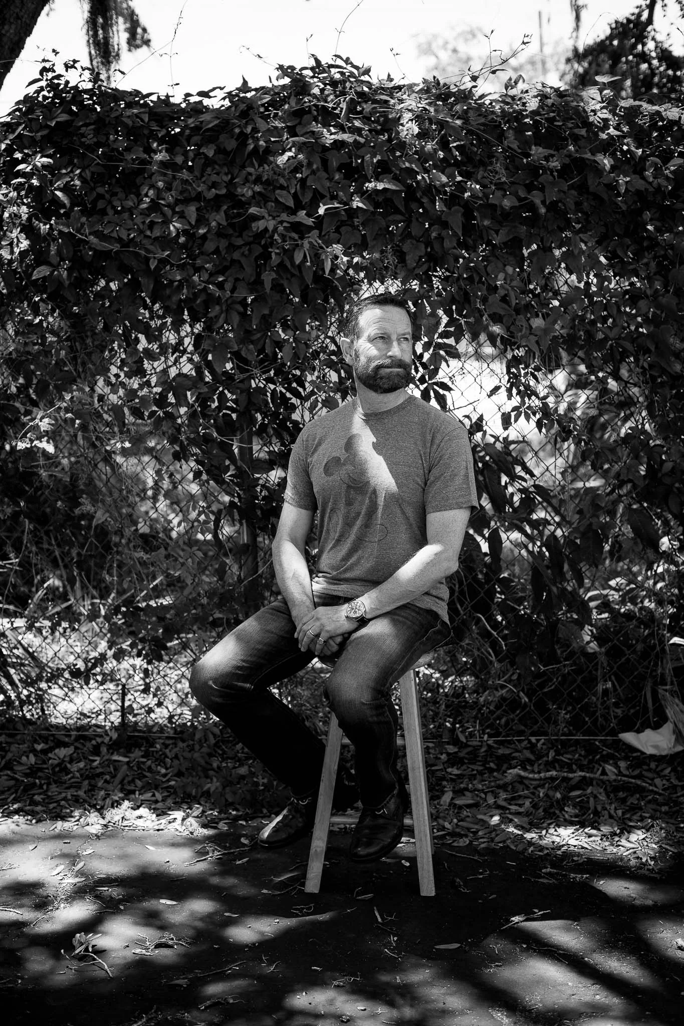  Black and white image of Duncan Wardle sitting outside on a stool in speckled light.  