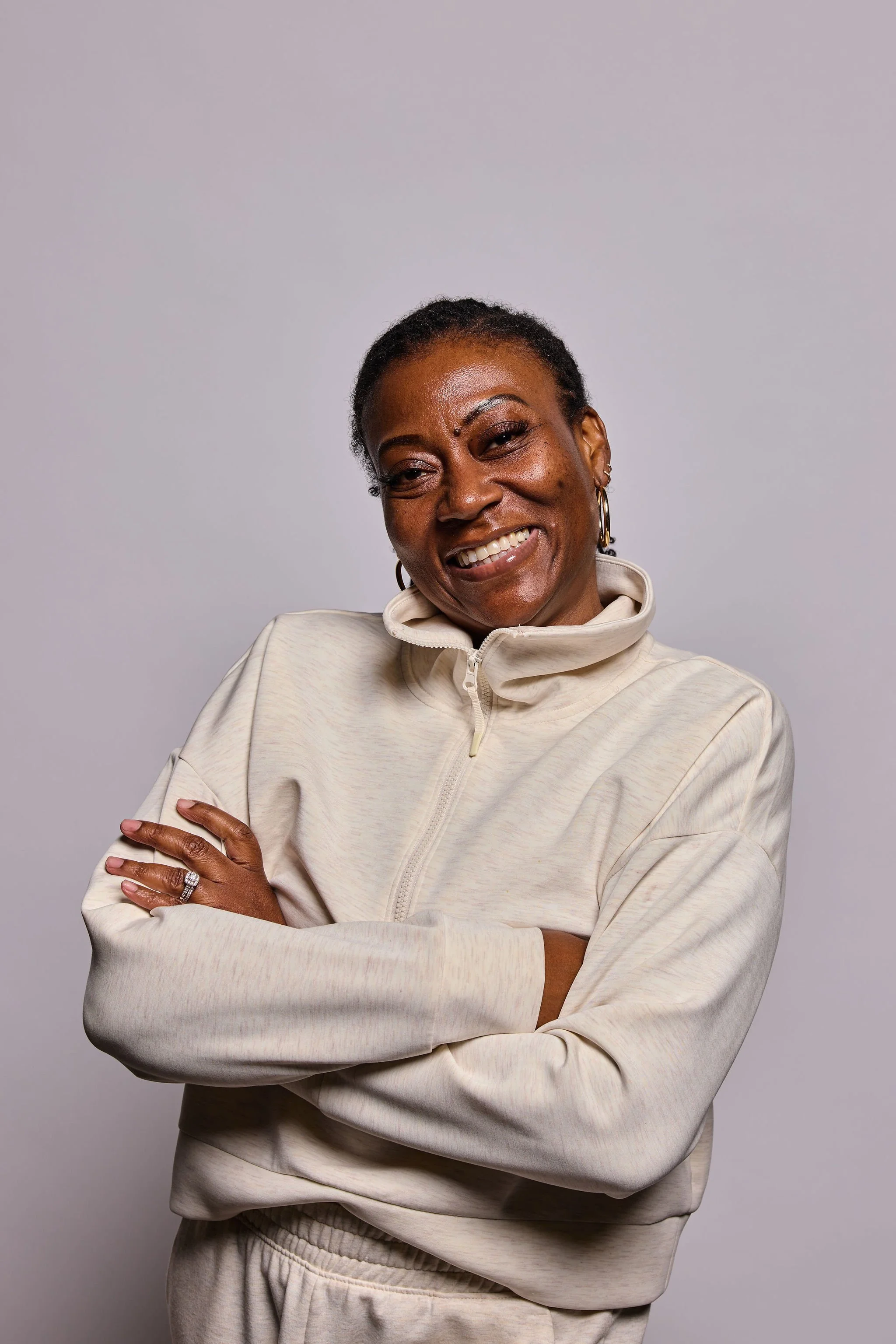 A woman with short hair and hoop earrings smiling, wearing a beige zip-up athletic jacket and pants, standing with her arms crossed in front of a plain gray background.
