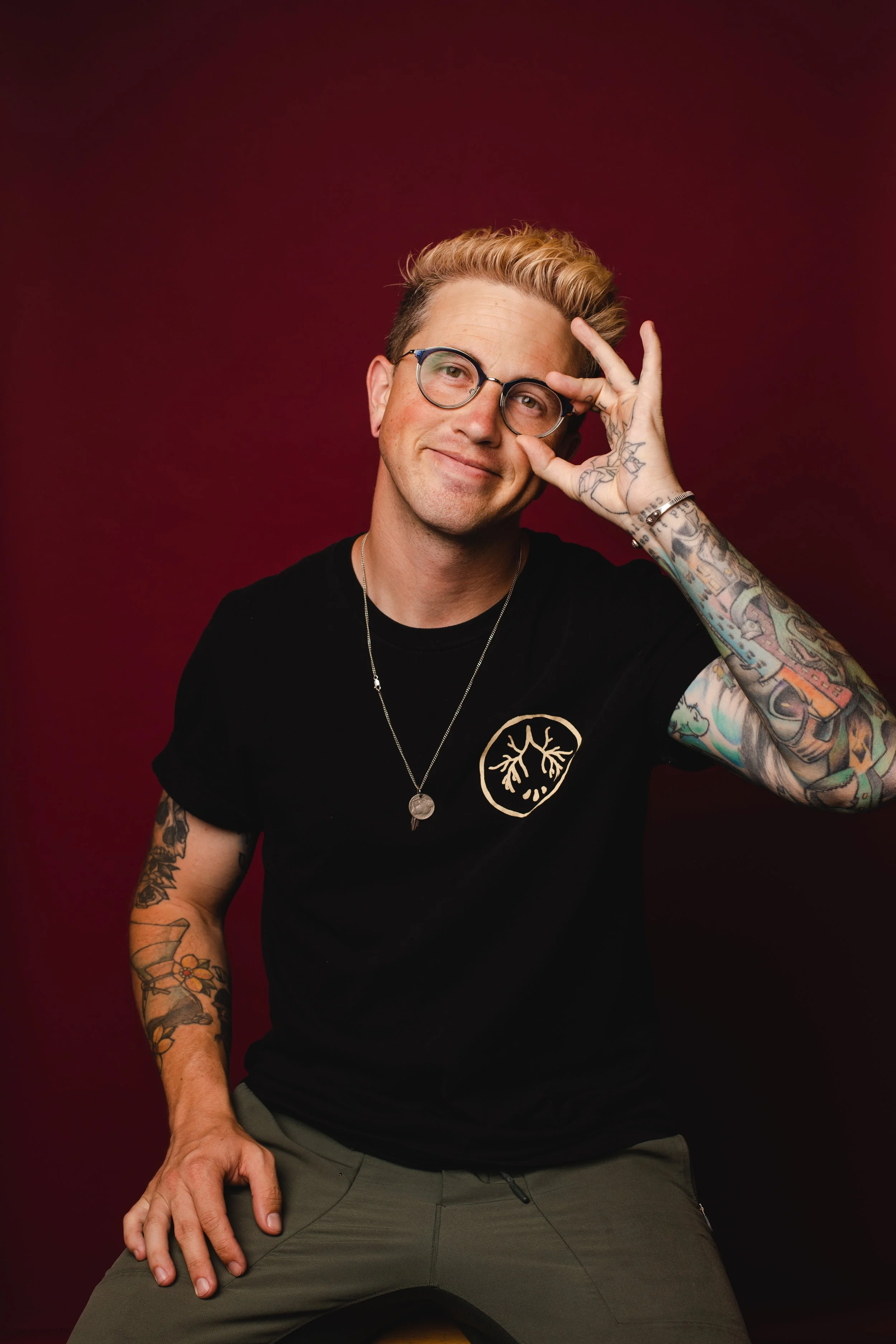 Portrait of a man with glasses, tattoos, blonde hair, smiling, sitting against a red background.