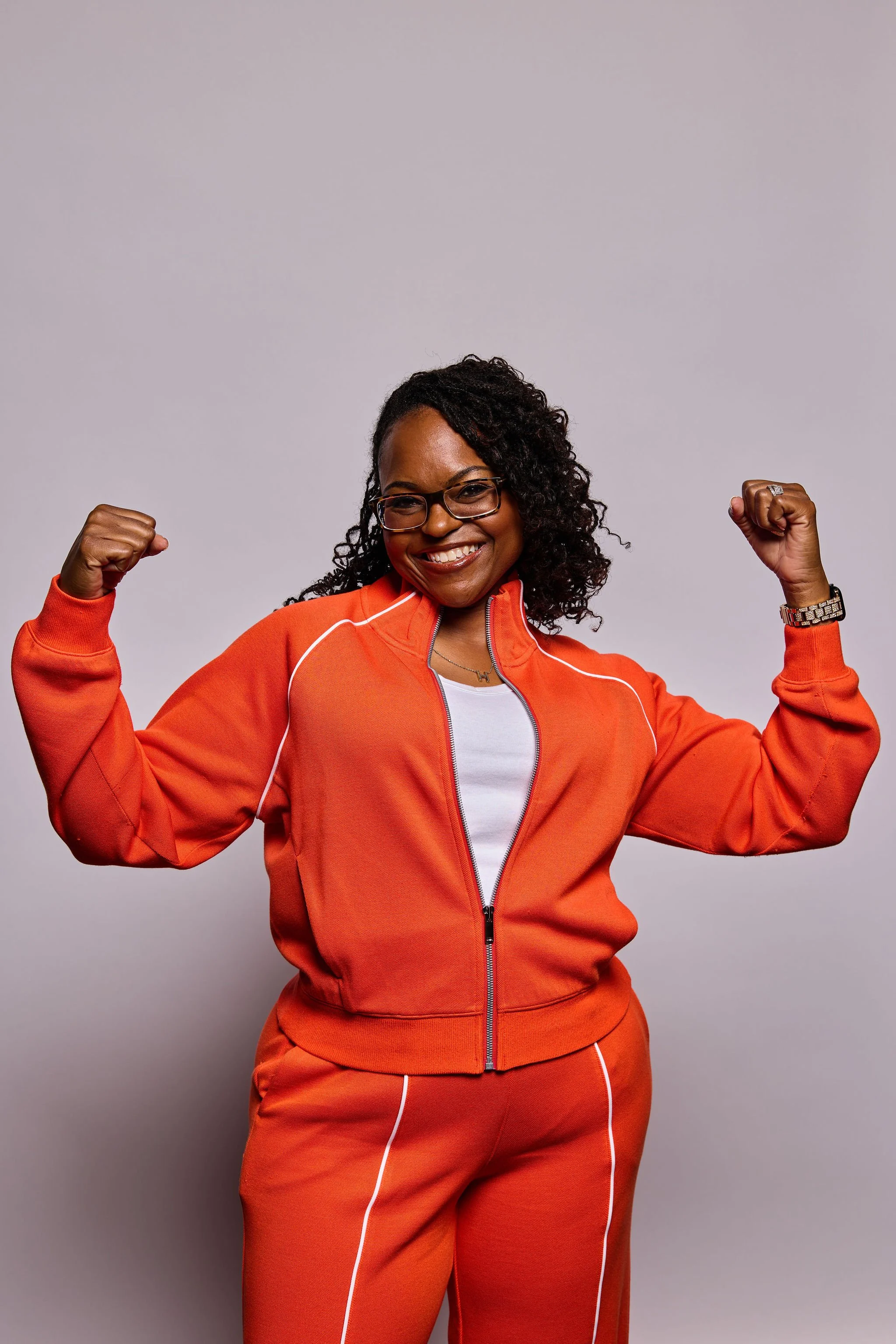 A woman with glasses and curly hair smiling and flexing her arms in a bright orange athletic tracksuit, standing against a plain light gray background.