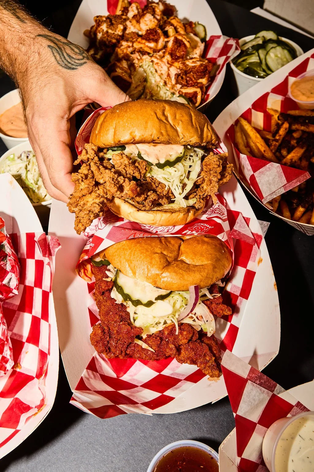 Close-up of a table with various food items, including fried chicken sandwiches with pickles and coleslaw, assorted fries, and side dishes, with a person's hand reaching towards the middle sandwich.