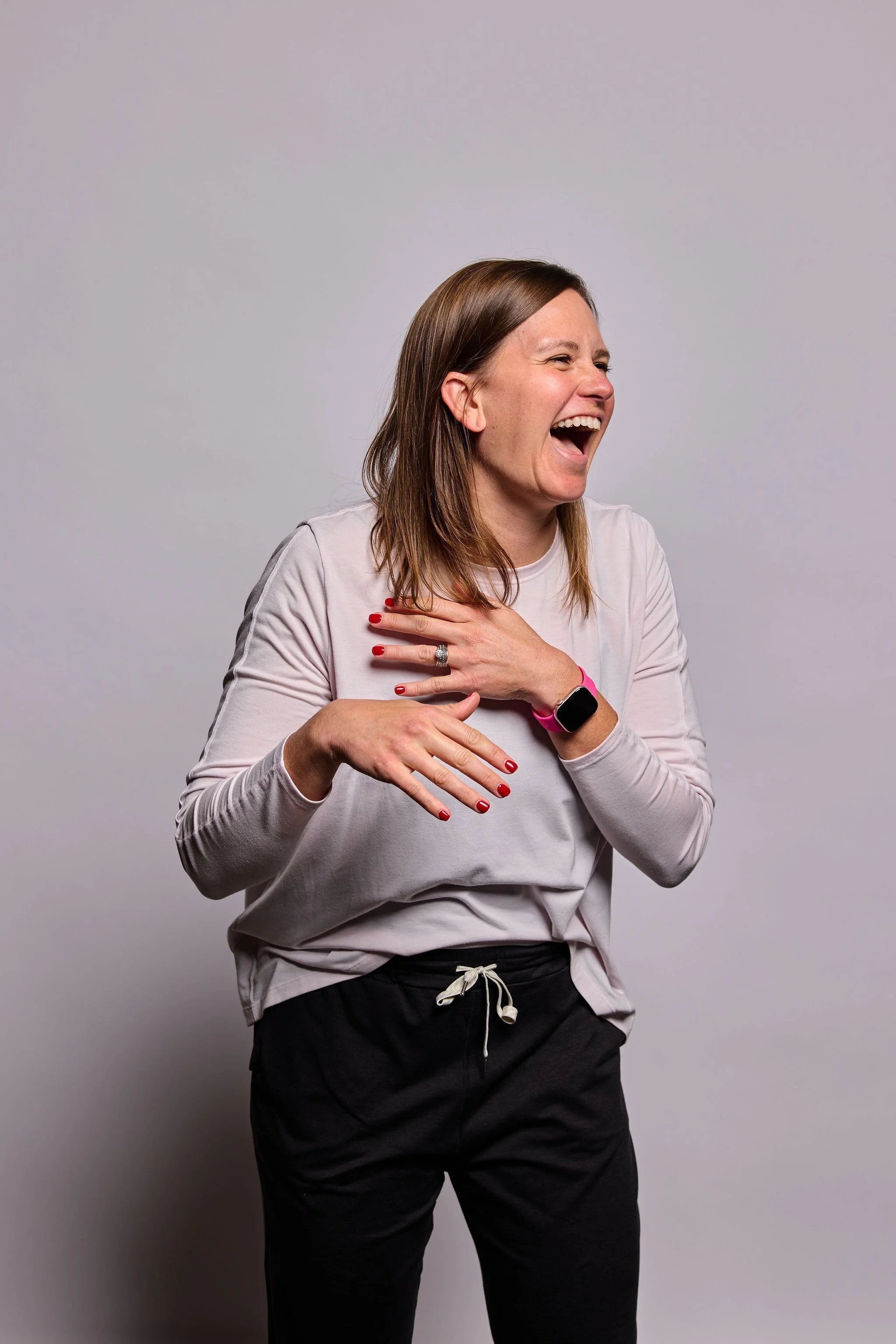 A woman with brown hair laughing and touching her chest with her right hand, wearing a light gray long-sleeve shirt, black pants with a white drawstring, a pink wristband, and a smartwatch, against a gray background.