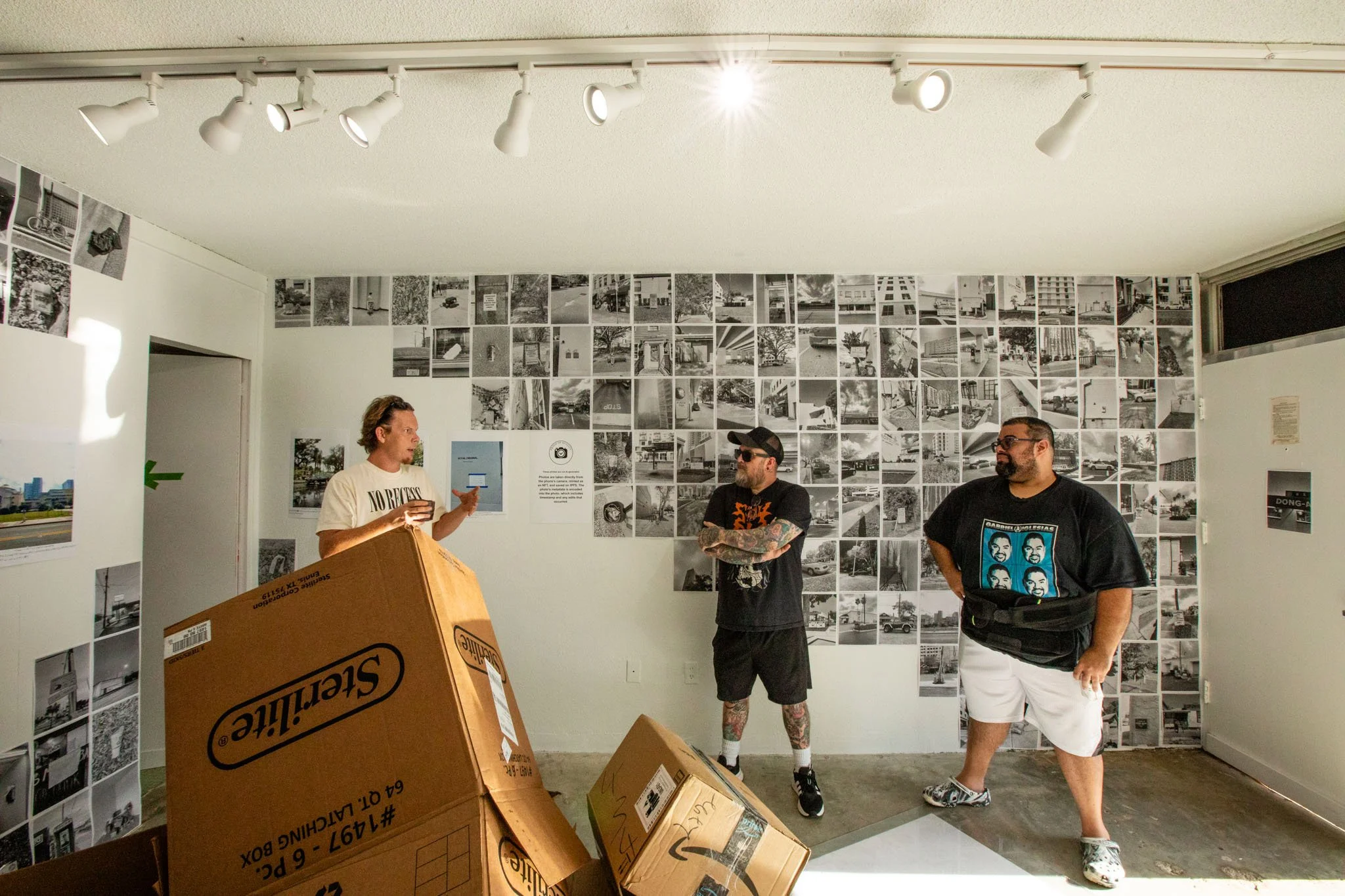 Three men in an art gallery with a wall covered in black and white photographs, boxes in the foreground, and ceiling spotlights illuminating the space.