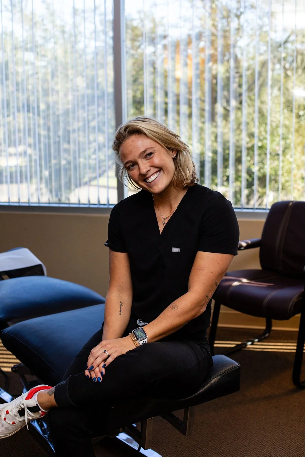 Smiling woman with blonde hair, tattoos, and a smartwatch sitting on an examination table in a medical office, with large windows and chairs in the background.
